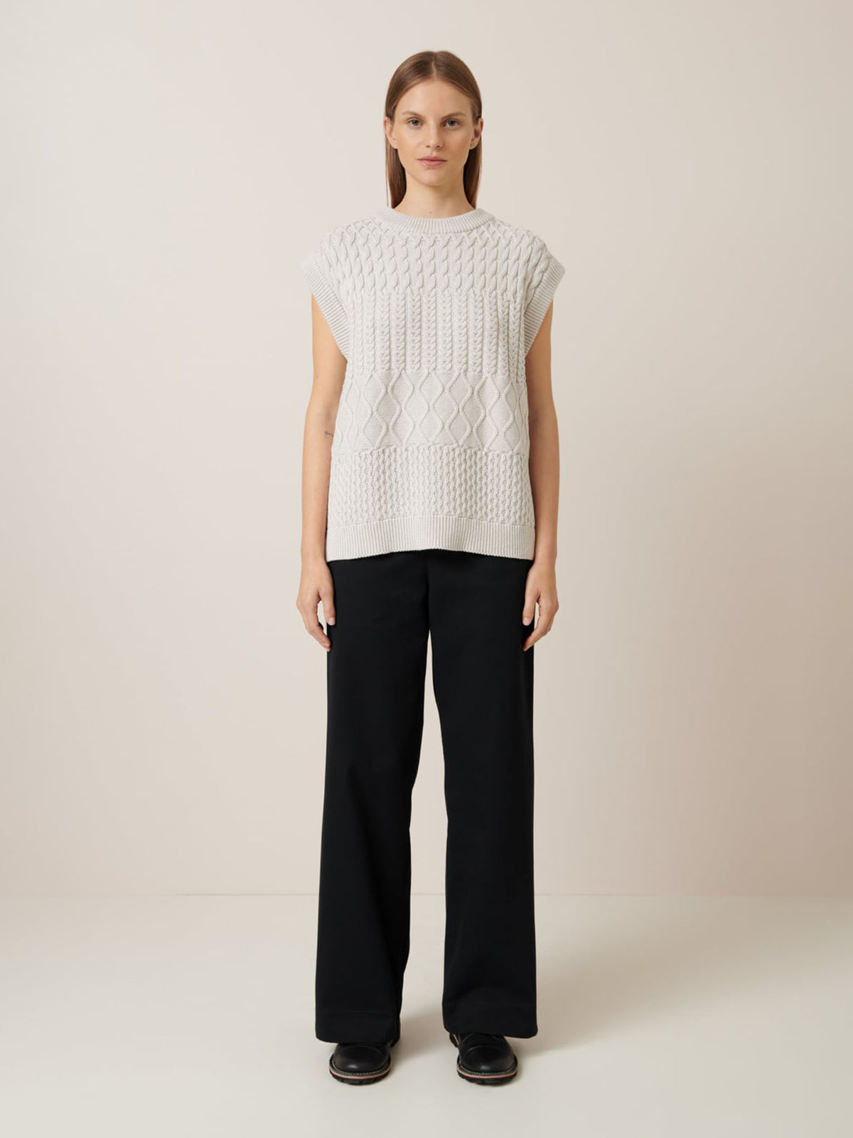 Person stands against a plain background, wearing an Aran Vest in Oat Melange and black wide-leg pants by Kowtow, arms by their sides.