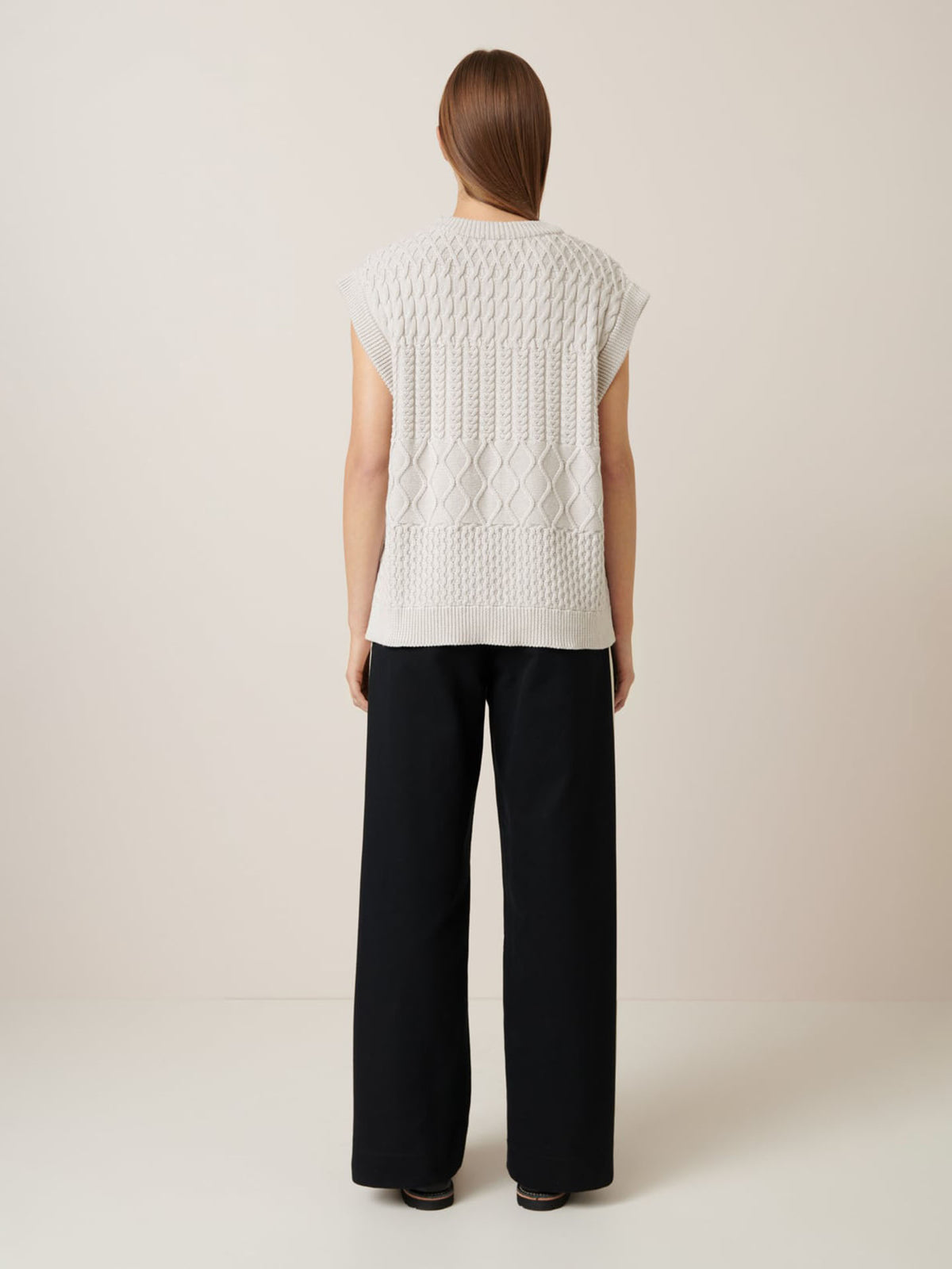 Against a plain backdrop, a person with long hair models the relaxed fit of Kowtow&#39;s Aran Vest in Oat Melange, effortlessly paired with black wide-leg pants.