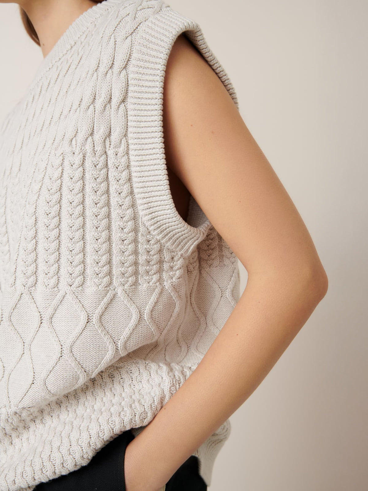 A person wears the Aran Vest – Oat Melange by Kowtow, a sleeveless, textured knit sweater vest. With its relaxed fit and neutral background, it offers comfort. Check the size guide for your perfect fit. Hands are tucked in pockets for a laid-back style.