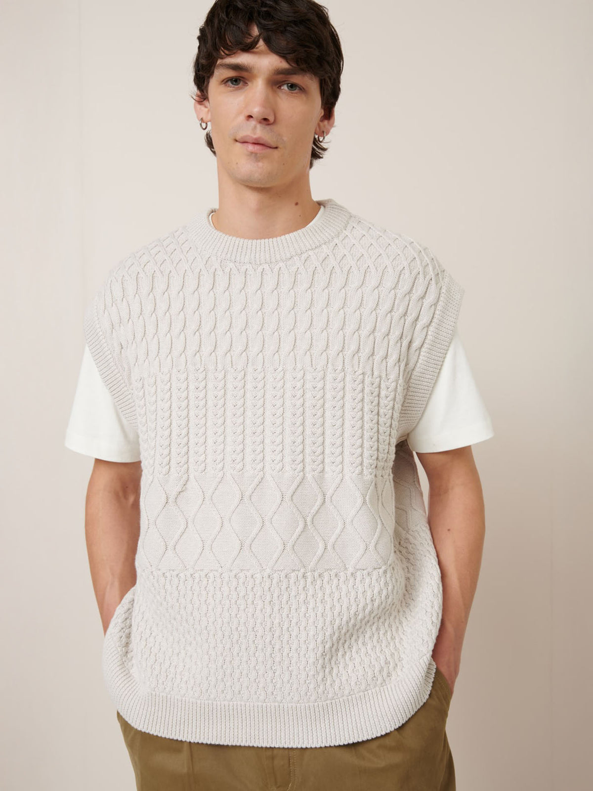 A person wears Kowtow&#39;s Aran Vest in Oat Melange over a white T-shirt with tan pants, highlighting the brand&#39;s relaxed fit style against a plain background.