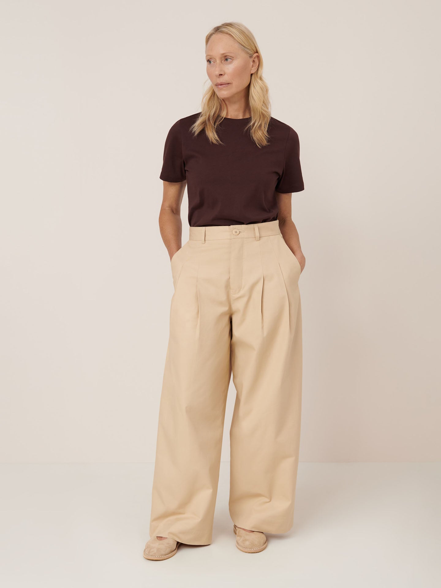 A woman stands against a plain background wearing a dark brown short-sleeve shirt, Kowtow Arch Pant – Dune (a relaxed fit, high-rise beige wide-leg pant), and beige shoes, her hands in her pockets.