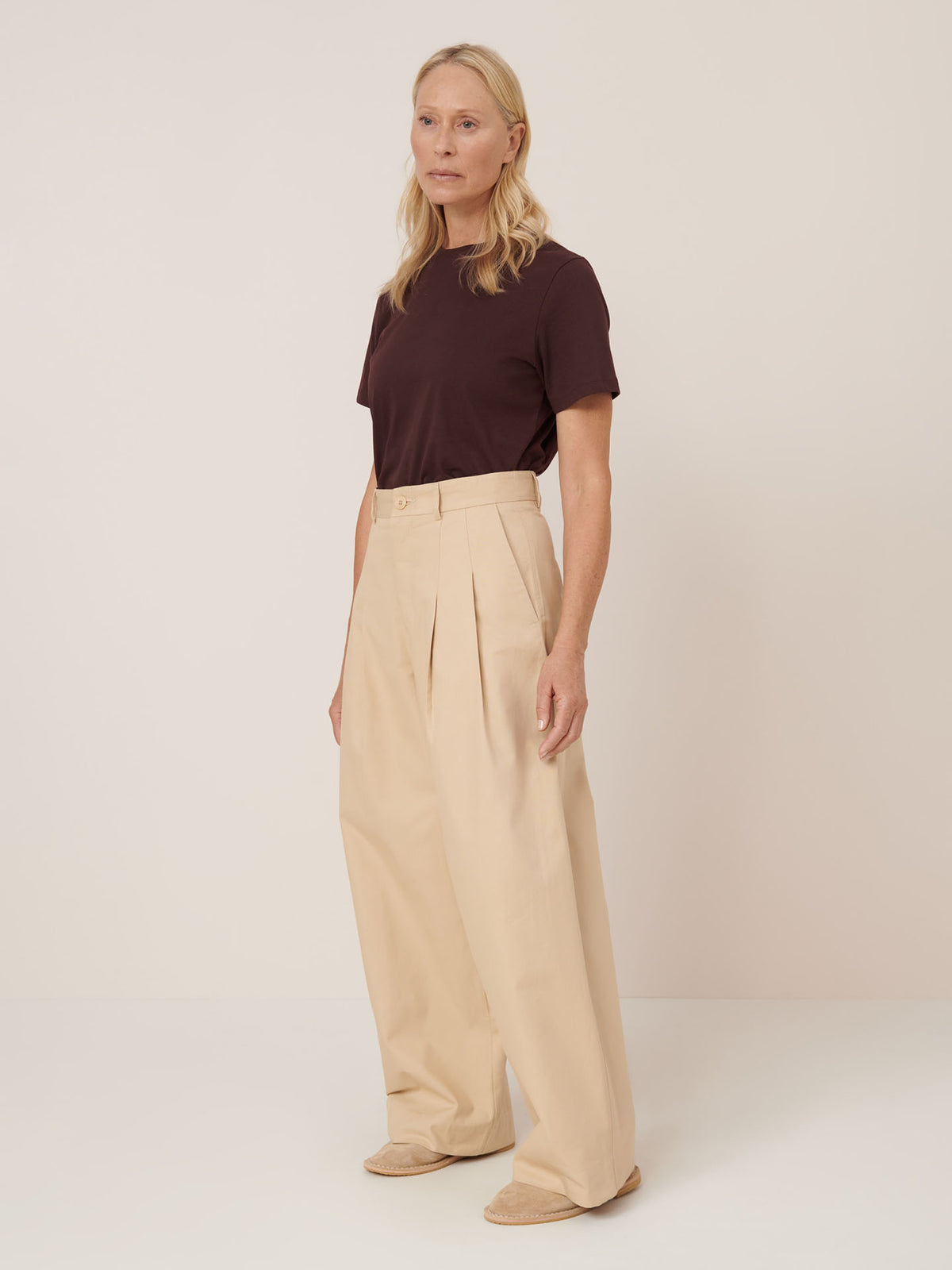 A person stands against a plain background wearing a dark brown t-shirt, Kowtow Arch Pant – Dune (relaxed fit, high-rise waist, beige wide-leg), and light-colored shoes.