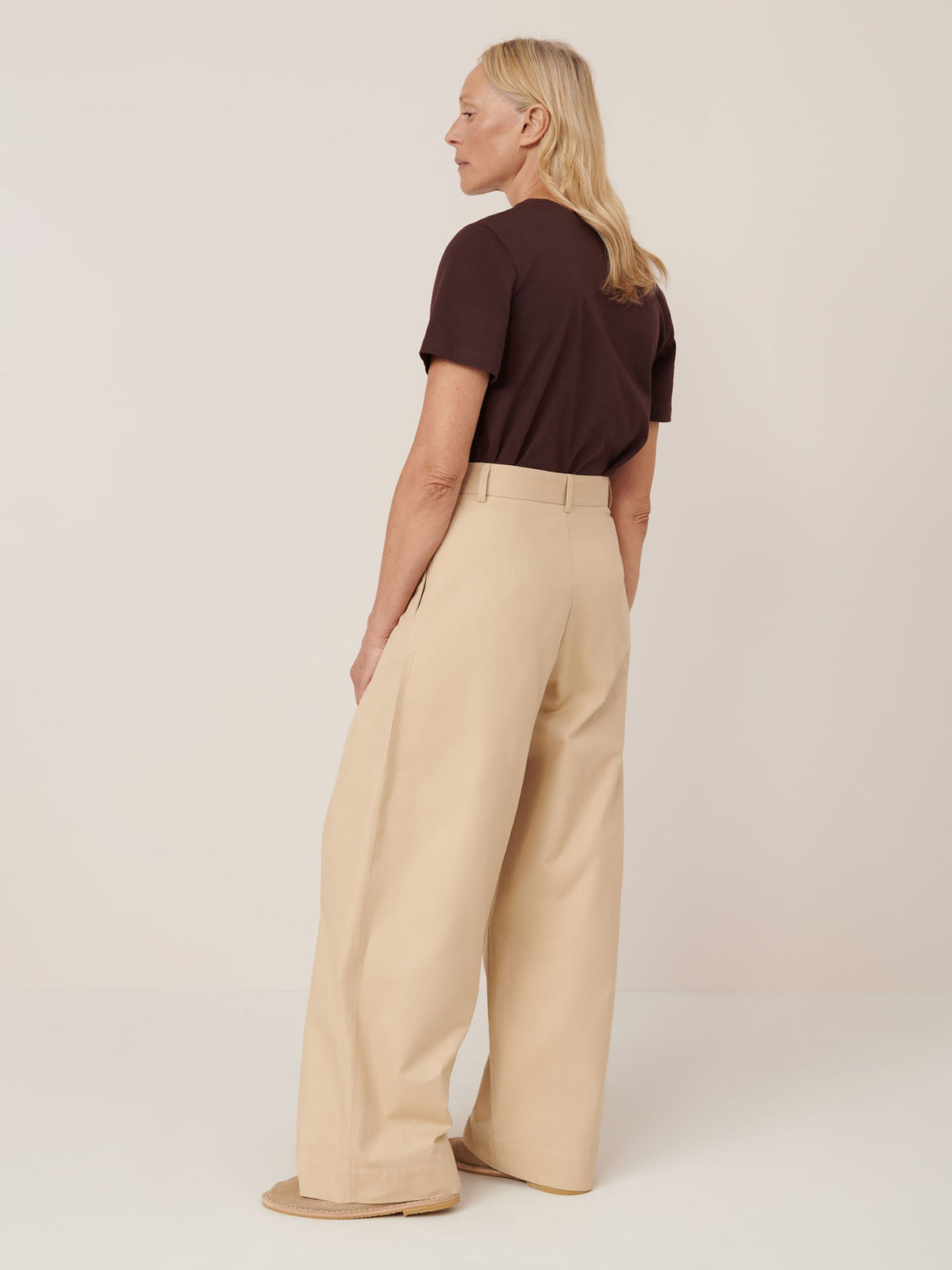 A woman stands sideways against a plain light background, wearing the Kowtow Arch Pant – Dune with a short-sleeve brown top and beige shoes.