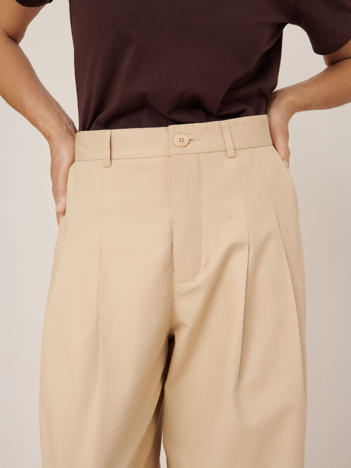 A person wears a dark brown t-shirt tucked into Kowtow’s Arch Pant – Dune, a high-rise, relaxed fit, beige pleated pant, with hands on hips.