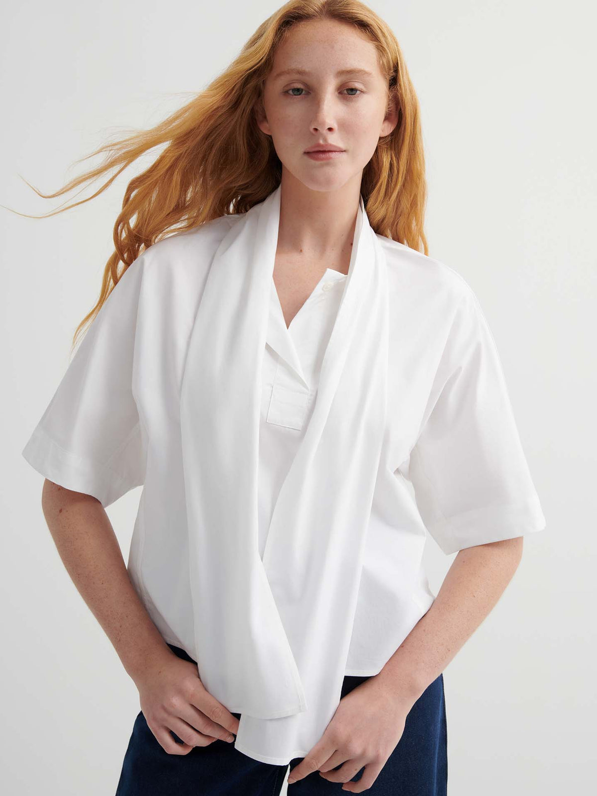 A person with long red hair wears a relaxed fit Ascot Top – White and dark pants, standing against a plain light background.
