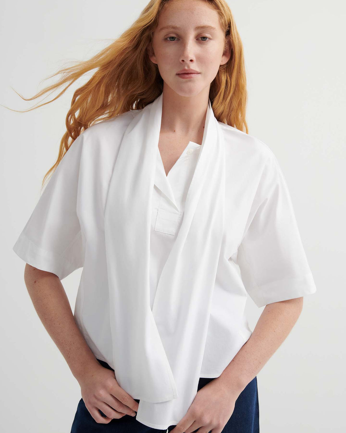 A person with long red hair wears a relaxed fit Ascot Top – White and dark pants, standing against a plain light background.