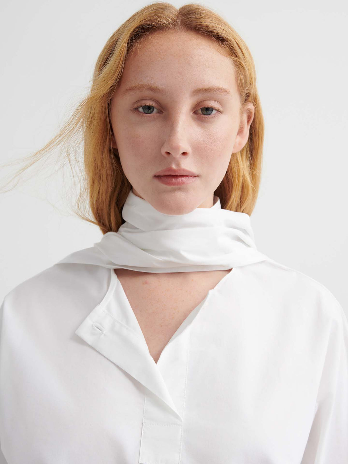 A person with light skin and red-blonde hair models the Ascot Top – White, styled in a relaxed fit with a scarf, against a plain light background.