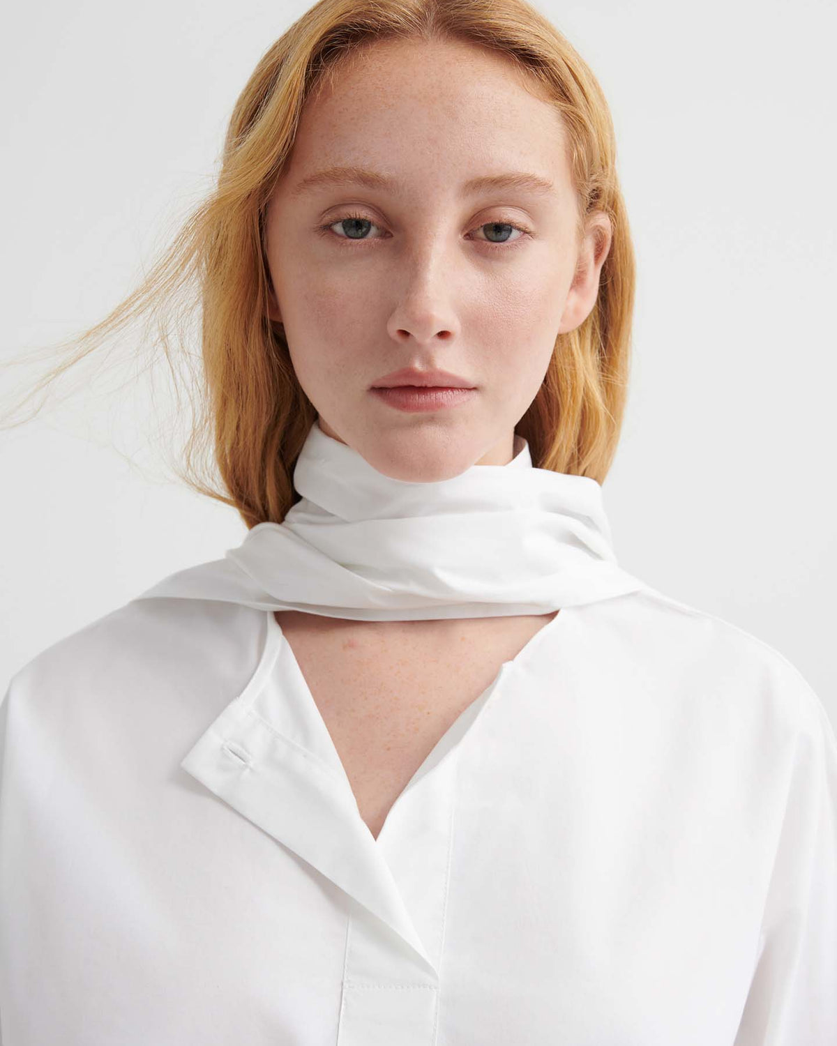 A person with light skin and red-blonde hair models the Ascot Top – White, styled in a relaxed fit with a scarf, against a plain light background.