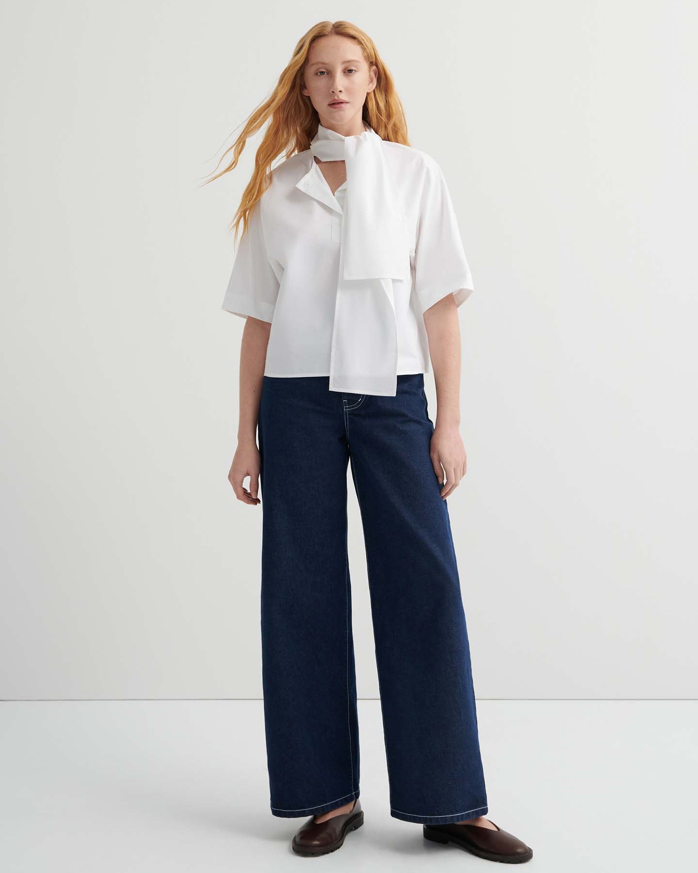 A woman with long red hair wears the Ascot Top – White with a scarf detail, paired with oversized wide-leg dark blue jeans and brown shoes, standing against a plain light background.