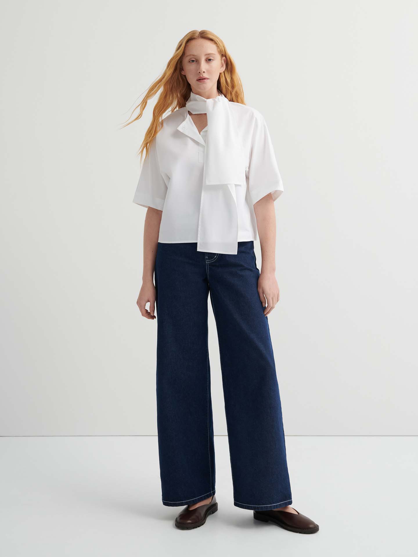 A woman with long red hair wears the Ascot Top – White with a scarf detail, paired with oversized wide-leg dark blue jeans and brown shoes, standing against a plain light background.