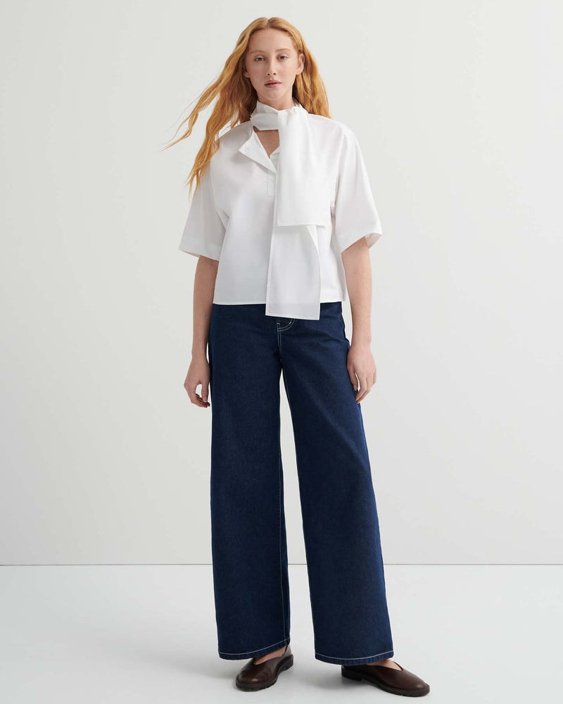 A woman with long red hair wears the Ascot Top – White with a scarf detail, paired with oversized wide-leg dark blue jeans and brown shoes, standing against a plain light background.
