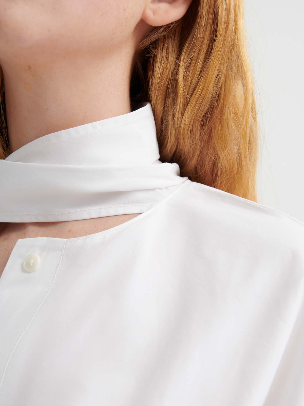 Close-up of a person with long, light brown hair wearing the Ascot Top – White, an oversized high-collared shirt with a button and scarf detail at the neck.