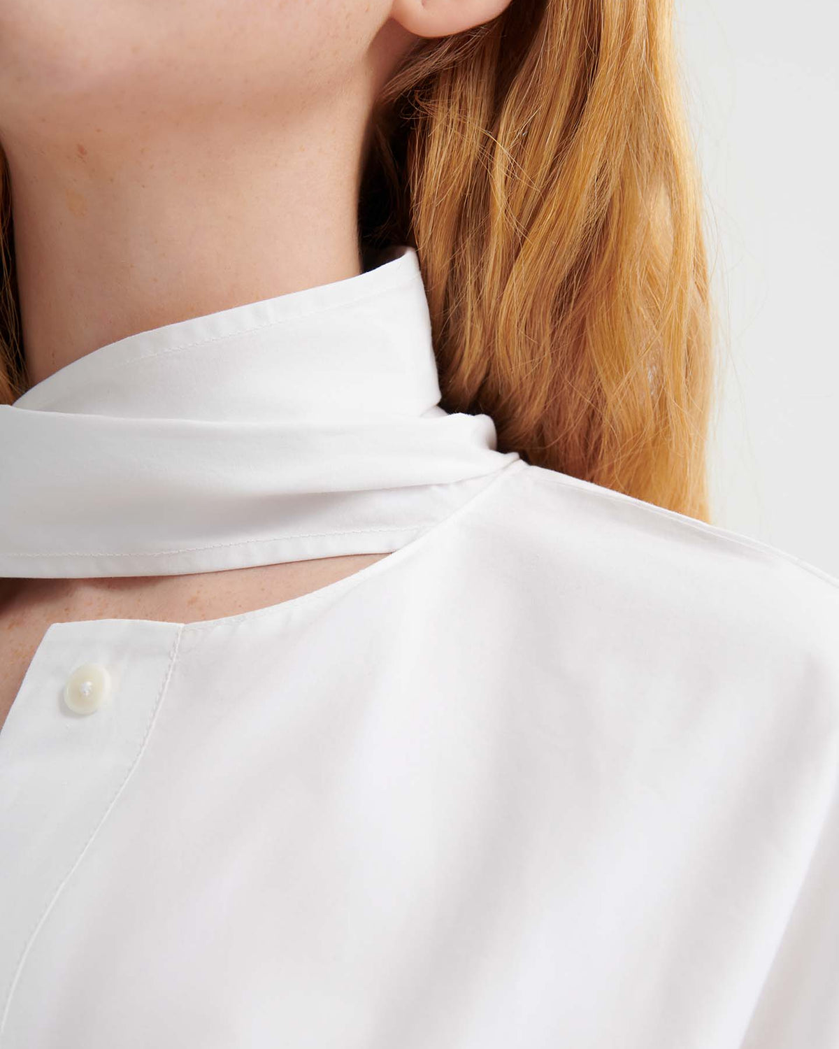Close-up of a person with long, light brown hair wearing the Ascot Top – White, an oversized high-collared shirt with a button and scarf detail at the neck.