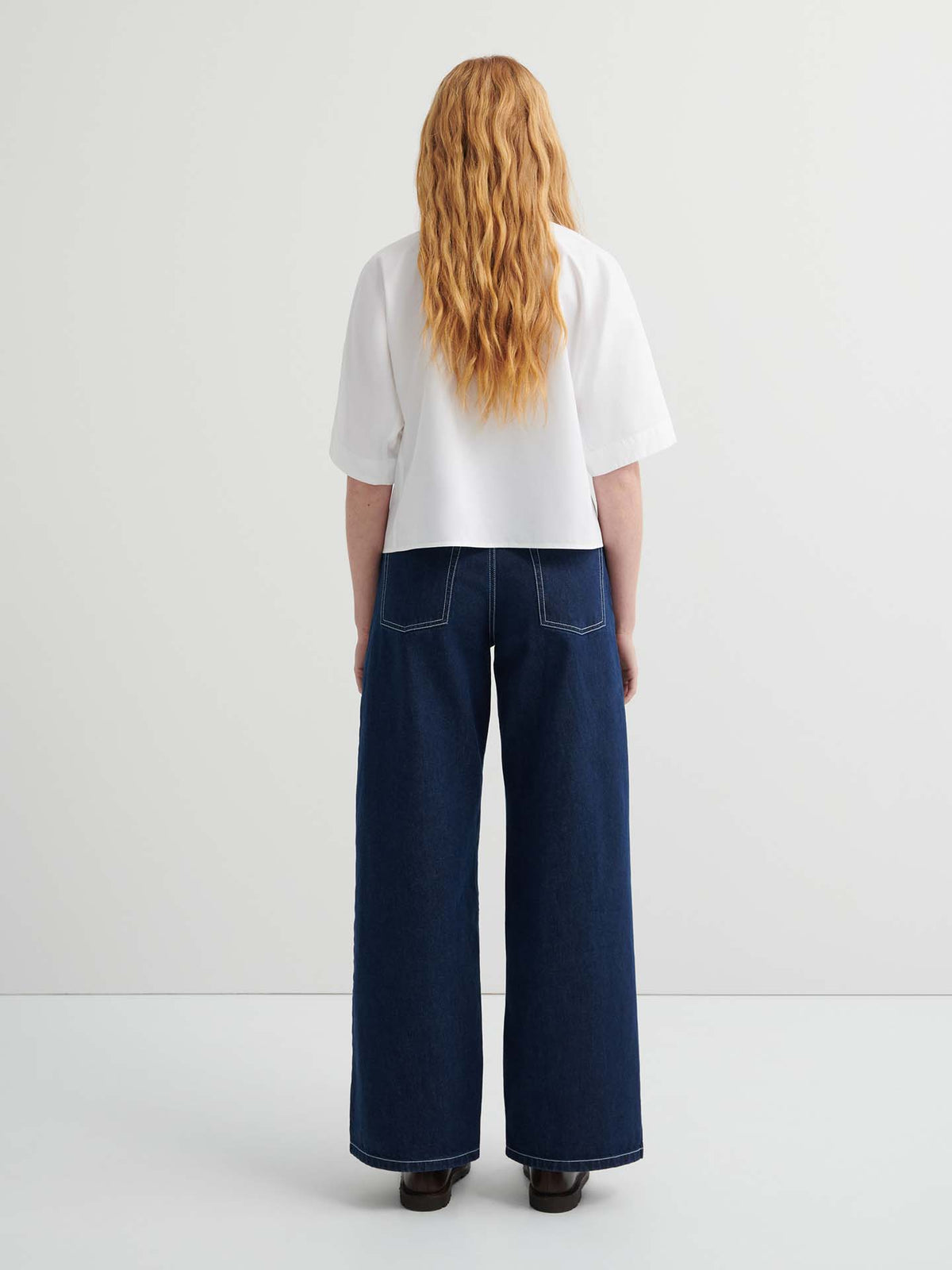 Person with long wavy hair wears the Ascot Top in white, a relaxed fit short-sleeve style, paired with wide-leg dark blue jeans, standing facing away against a plain light background.