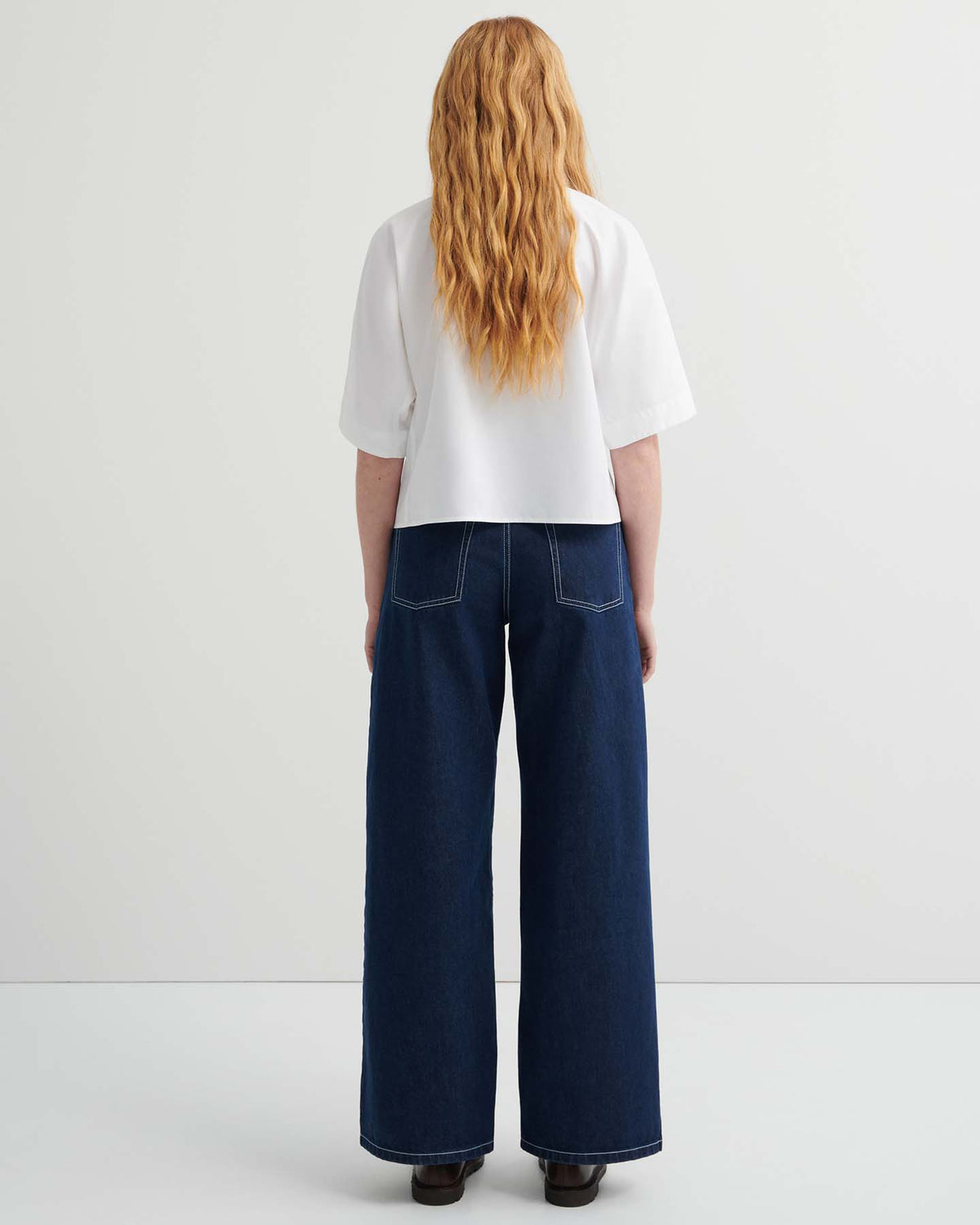 Person with long wavy hair wears the Ascot Top in white, a relaxed fit short-sleeve style, paired with wide-leg dark blue jeans, standing facing away against a plain light background.