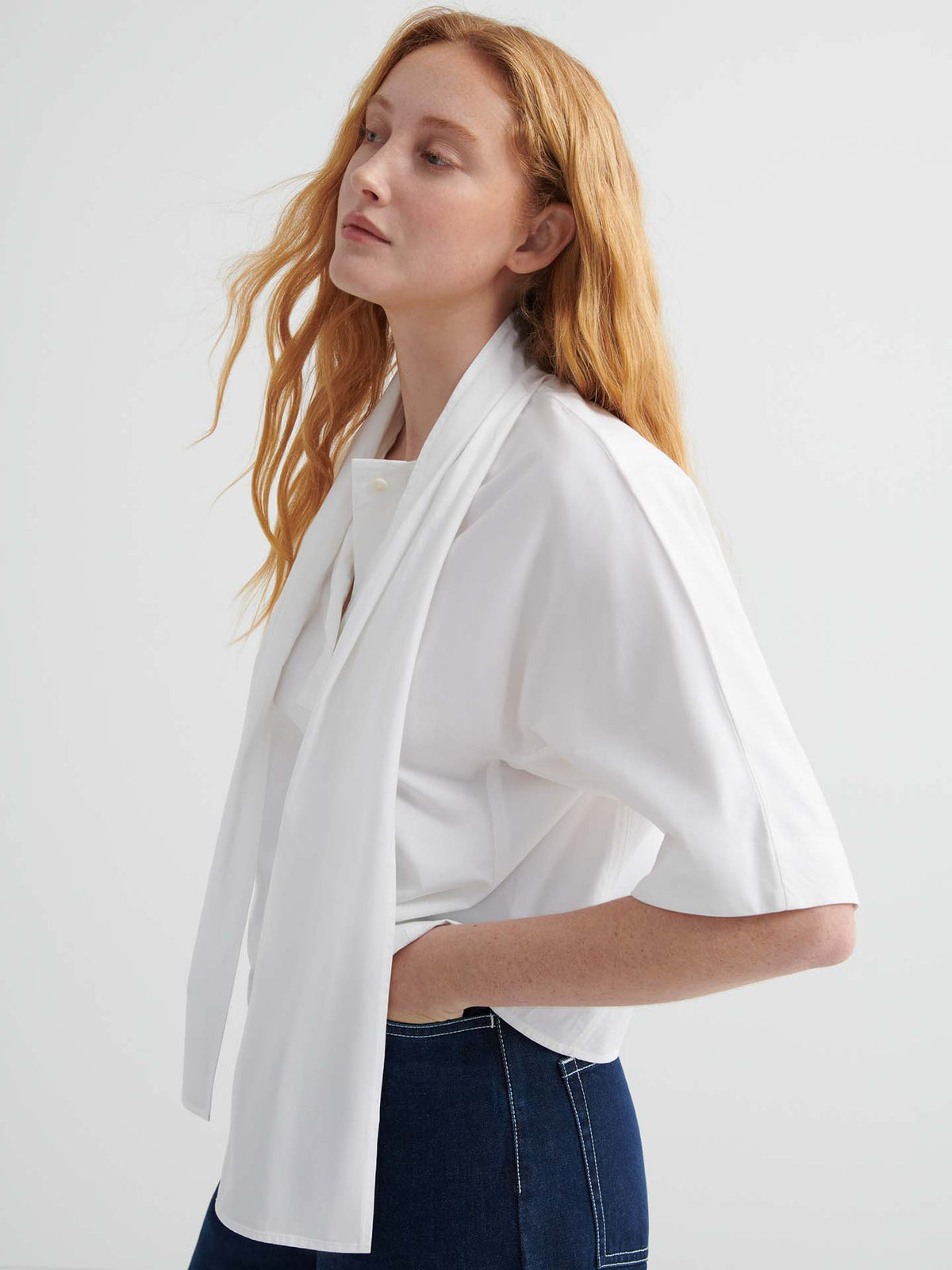 A person with long red hair wears the Ascot Top – White and dark jeans, standing against a plain light background.