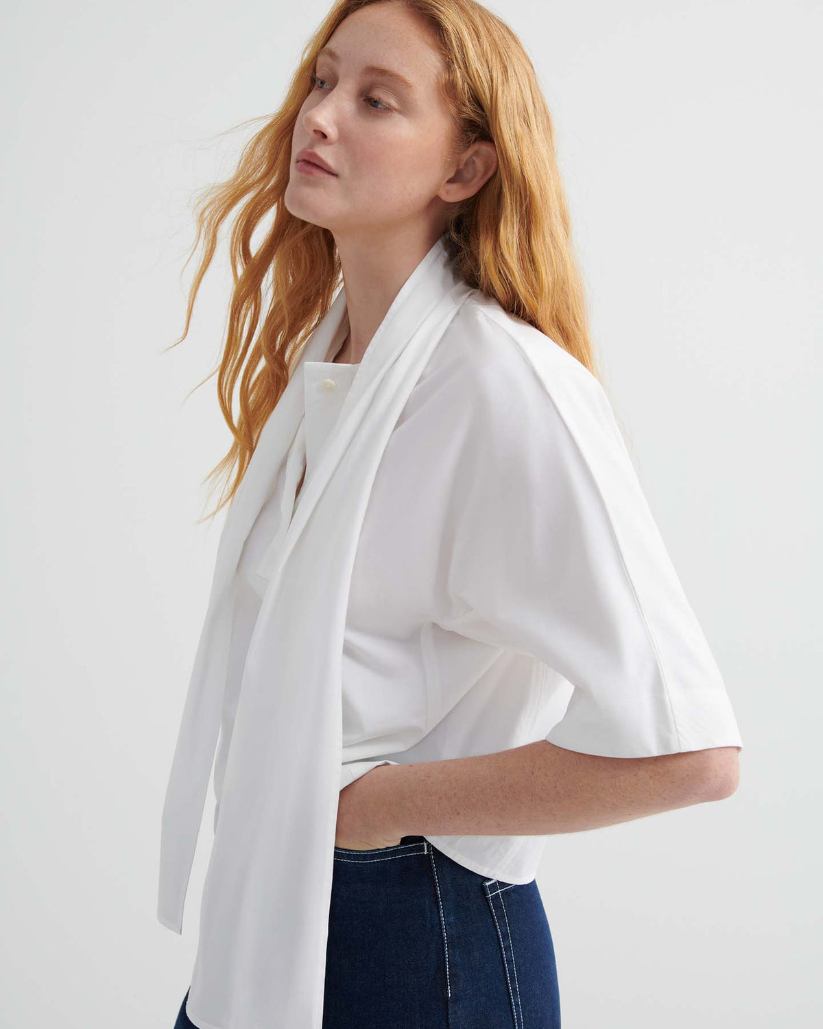 A person with long red hair wears the Ascot Top – White and dark jeans, standing against a plain light background.