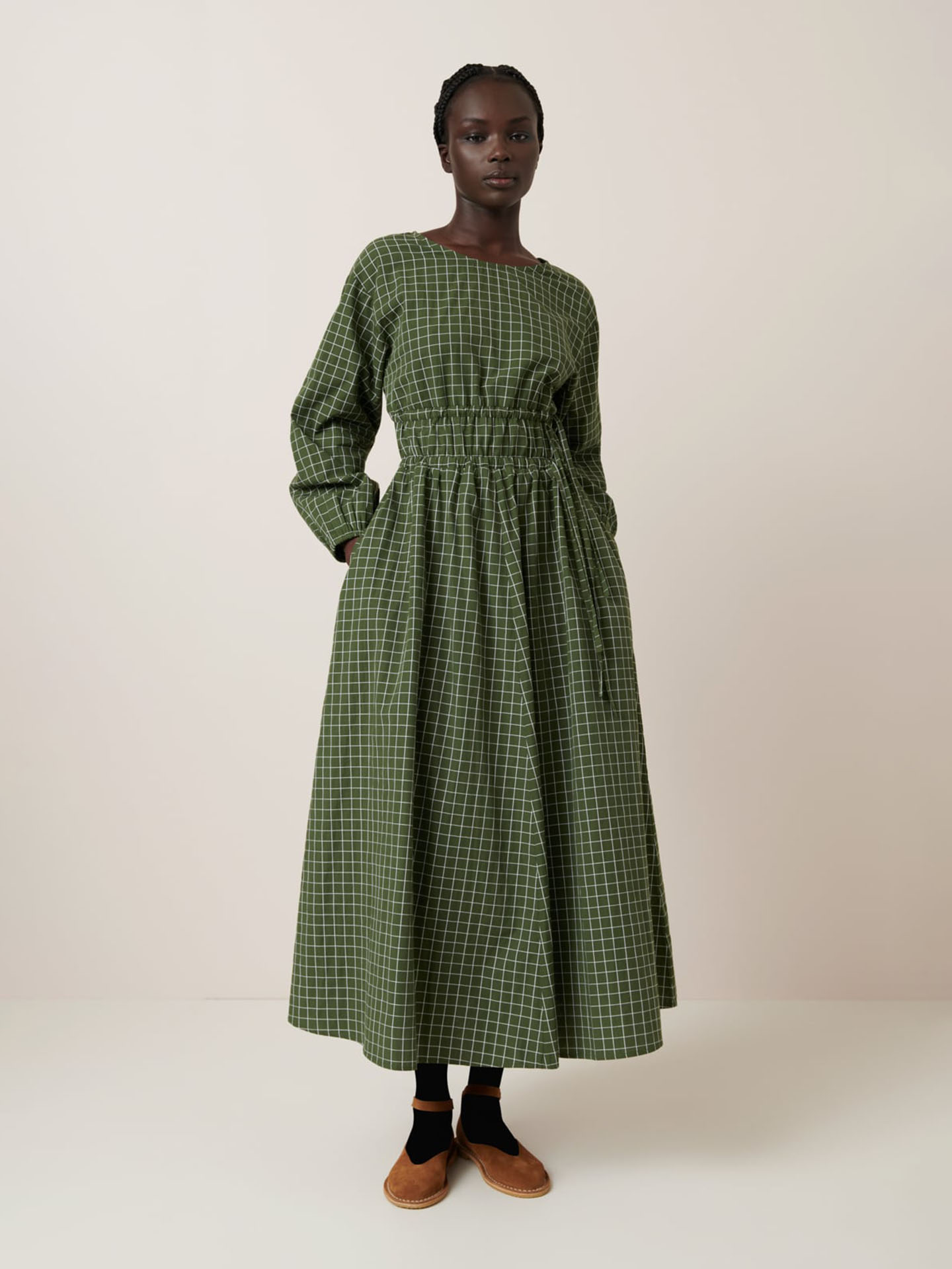 A person wears the Kowtow Aspen Dress – Forest Grid, a relaxed fit, long-sleeved green checkered dress with a gathered waist, paired with black socks and brown shoes. See size guide for measurements.
