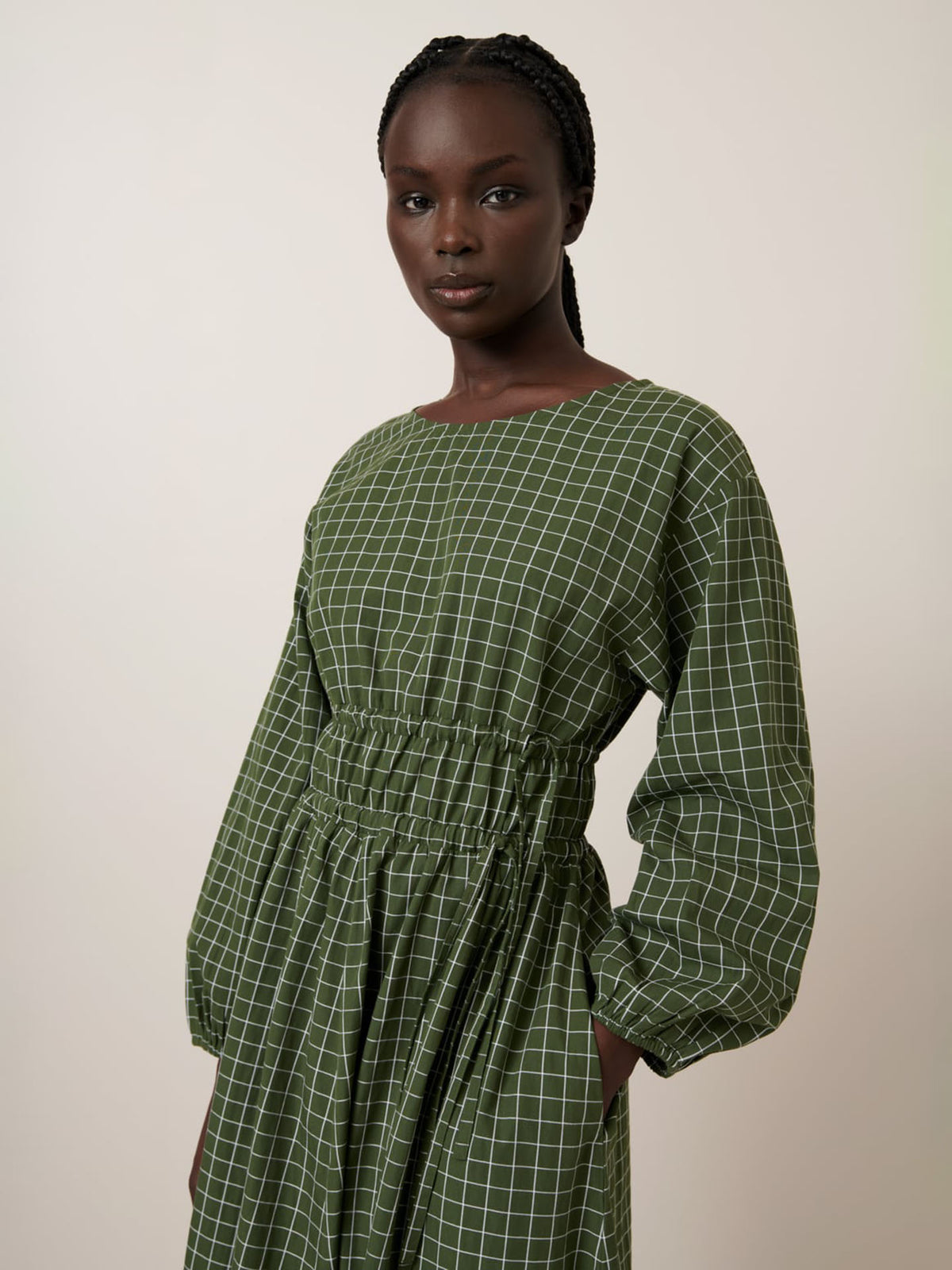 A woman wears the Kowtow Aspen Dress – Forest Grid, a long-sleeved green dress with a white grid pattern, elastic waist, and relaxed fit. See our size guide and garment measurements for more details.