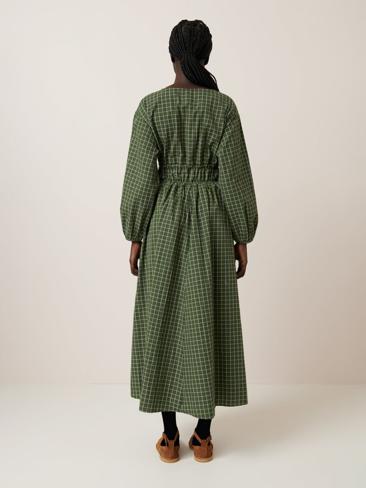 A person with braided hair faces away, wearing the Kowtow Aspen Dress – Forest Grid, a relaxed fit, long-sleeved green checkered dress with a cinched waist, paired with black leggings and tan platform sandals in a neutral setting.