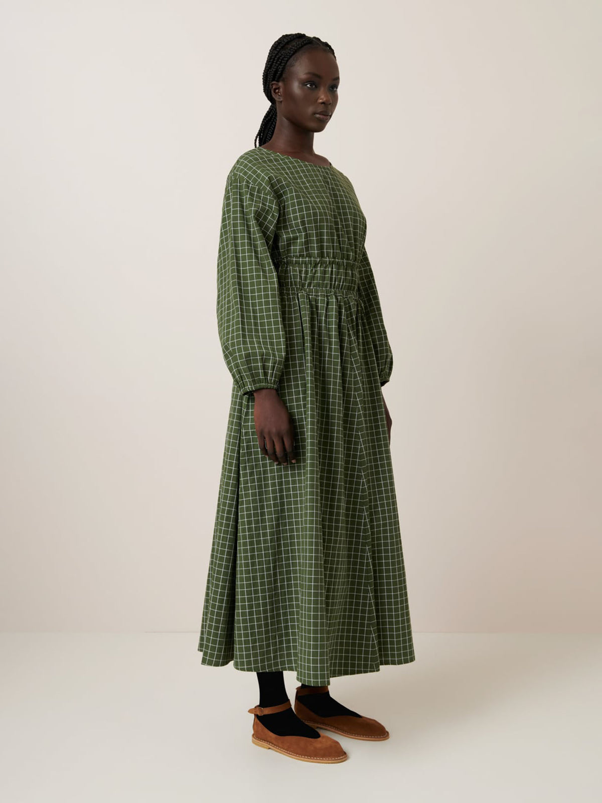 A person stands in profile wearing Kowtow’s relaxed fit Aspen Dress – Forest Grid with a cinched waist, paired with black leggings and brown flats, against a plain light background.