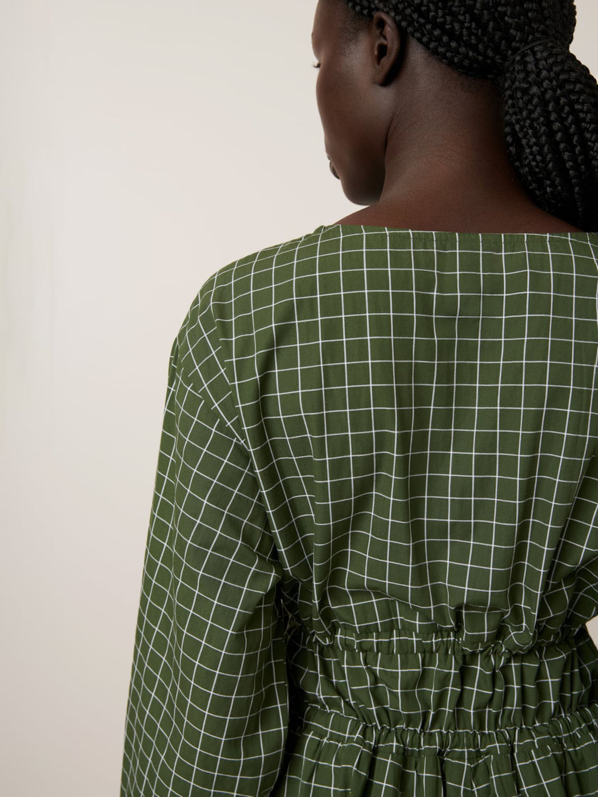 A person with braided hair is seen from behind wearing the Kowtow Aspen Dress – Forest Grid, featuring a relaxed fit, white grid pattern, and an elastic waistband.