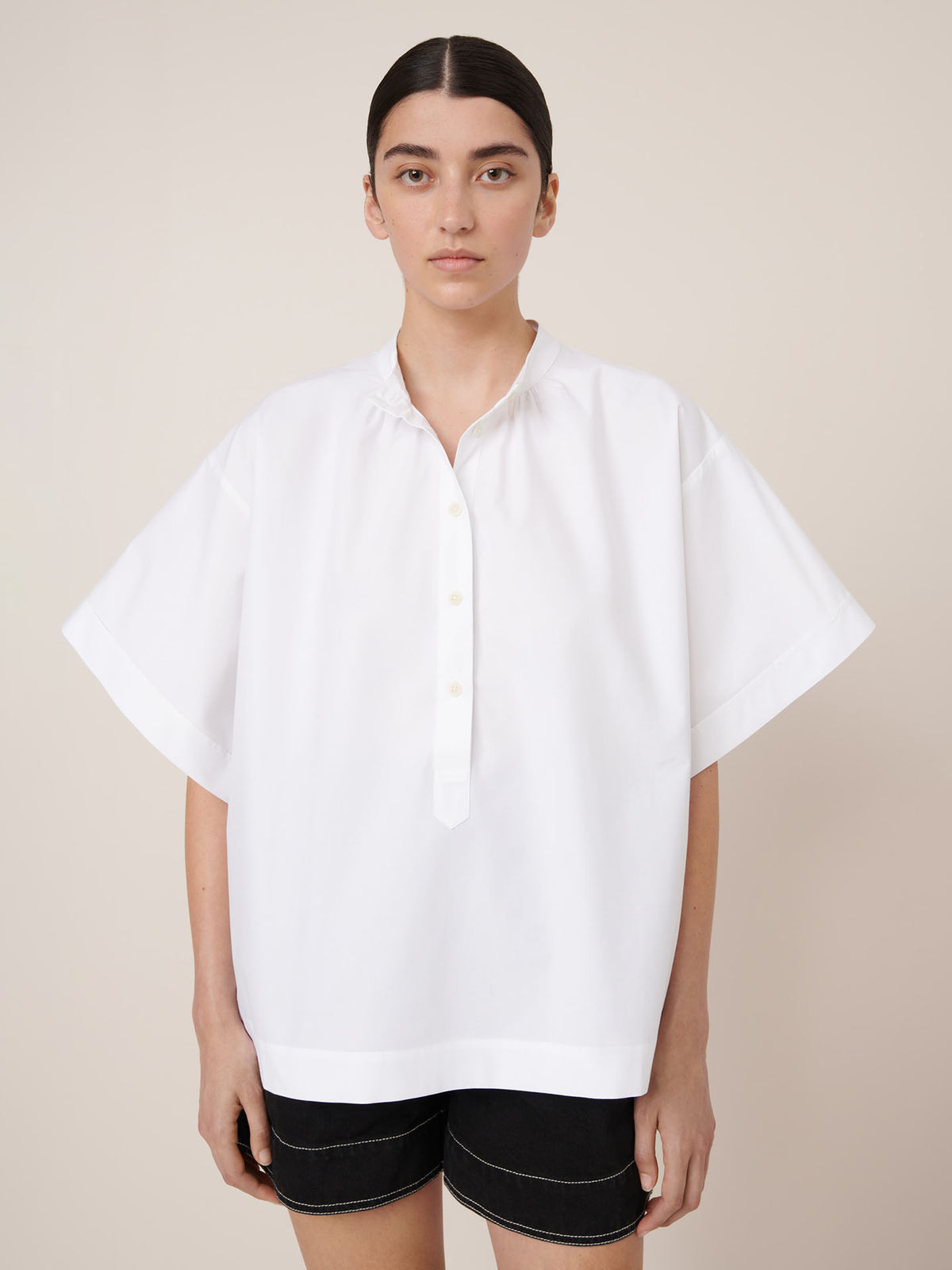 A person stands against a plain background, wearing the Kowtow Atelier Shirt in white—an oversized, loose short-sleeve button-up—paired with black shorts and looking directly at the camera.