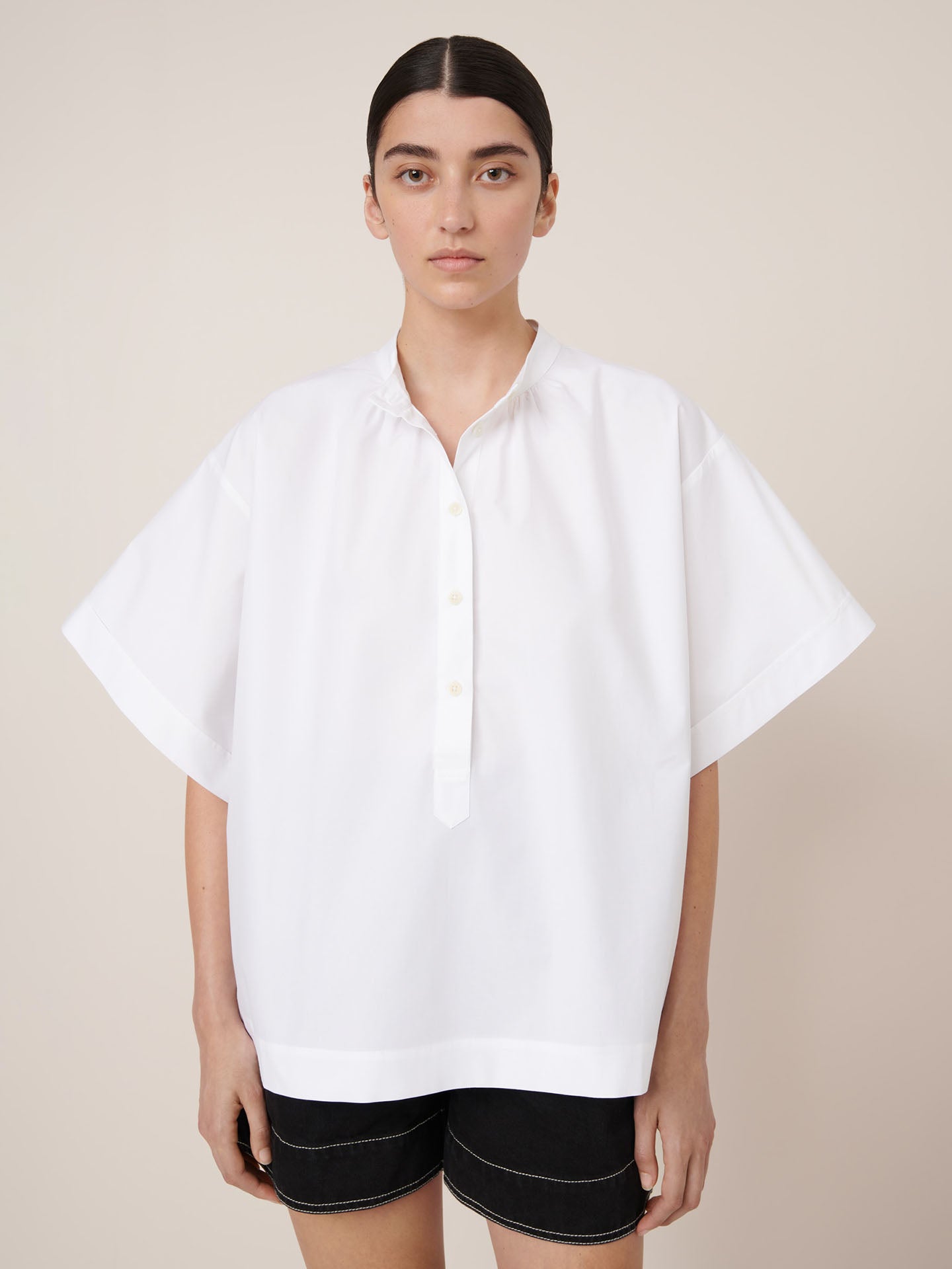 A person stands against a plain background, wearing the Kowtow Atelier Shirt in white—an oversized, loose short-sleeve button-up—paired with black shorts and looking directly at the camera.