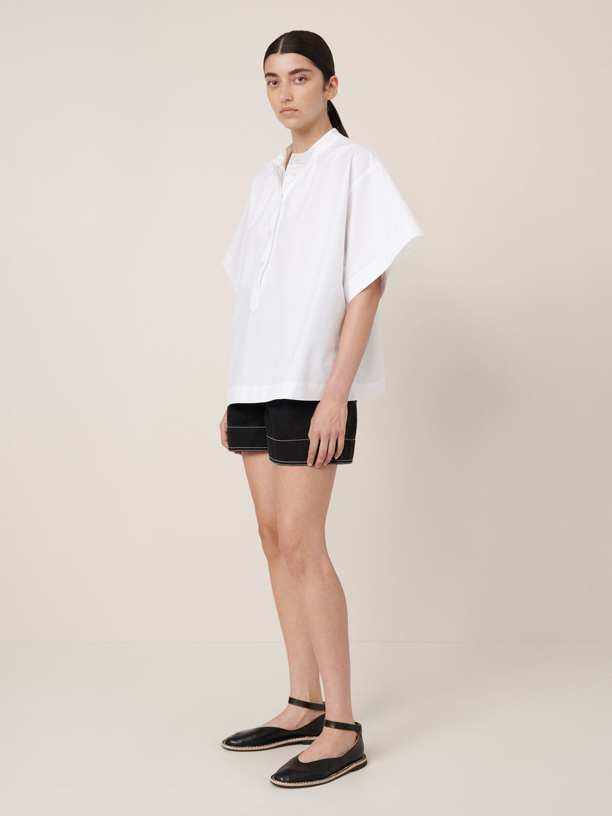 A woman stands against a plain background wearing the Kowtow Atelier Shirt in white—a loose, oversized short-sleeve top paired with black shorts and black ankle-strap flats, evoking Kowtow&#39;s signature boxy aesthetic.