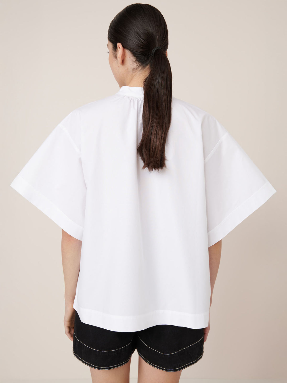 A person with long brown hair in a low ponytail wears Kowtow’s Atelier Shirt in white and black shorts, shown from the back against a plain background, showcasing the brand’s signature oversized fit.