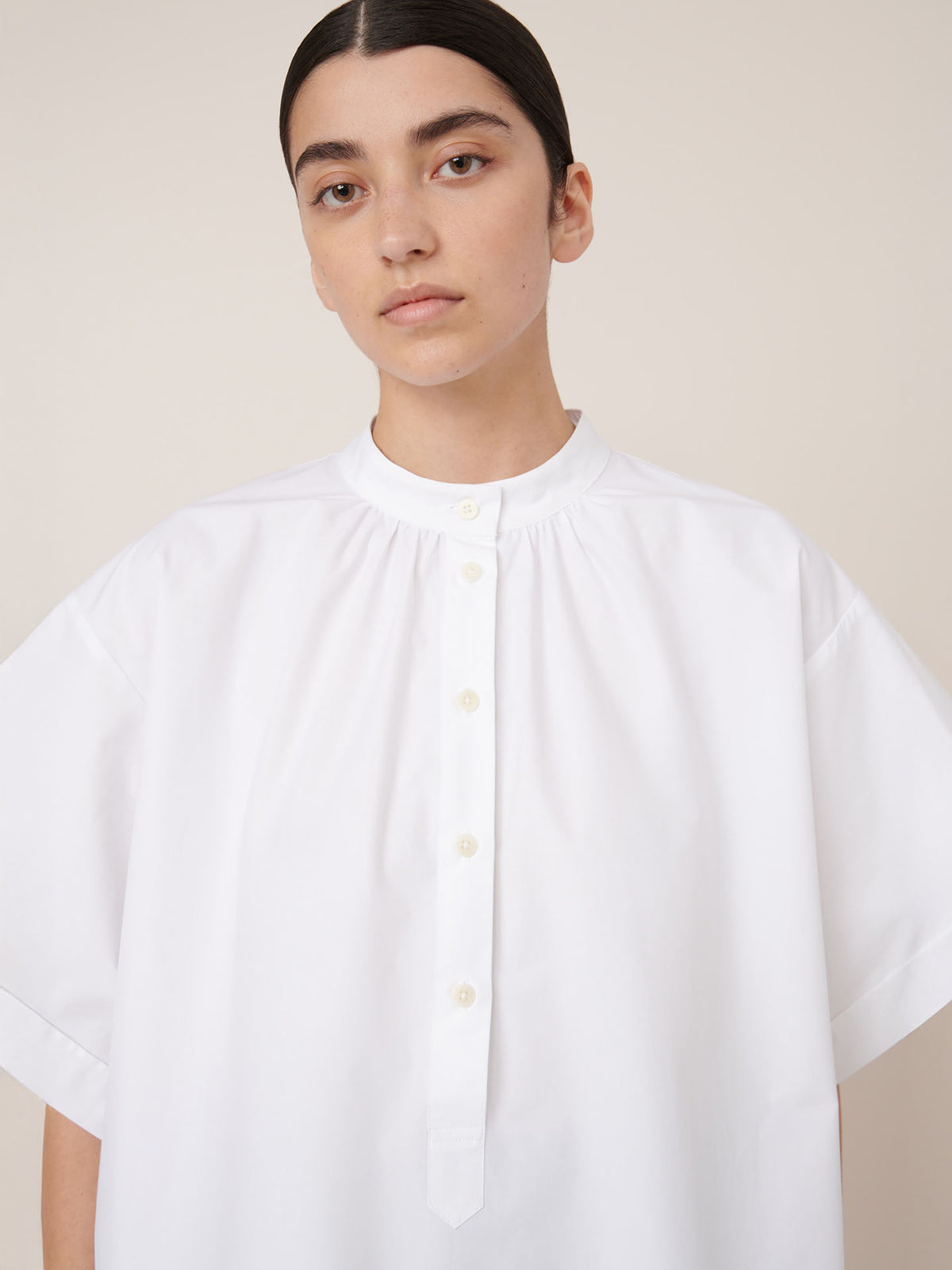 A person with dark hair pulled back wears the Kowtow Atelier Shirt – White, a loose-fitting, short-sleeve, oversized button-up, and looks directly at the camera.