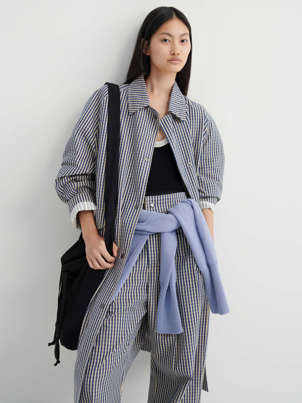 A person stands against a white wall wearing a striped jacket and pants, a black tank top, and a light blue sweater tied at the waist, carrying a black bag styled with the Atelier Trench – Landscape Check for a relaxed trench coat look.