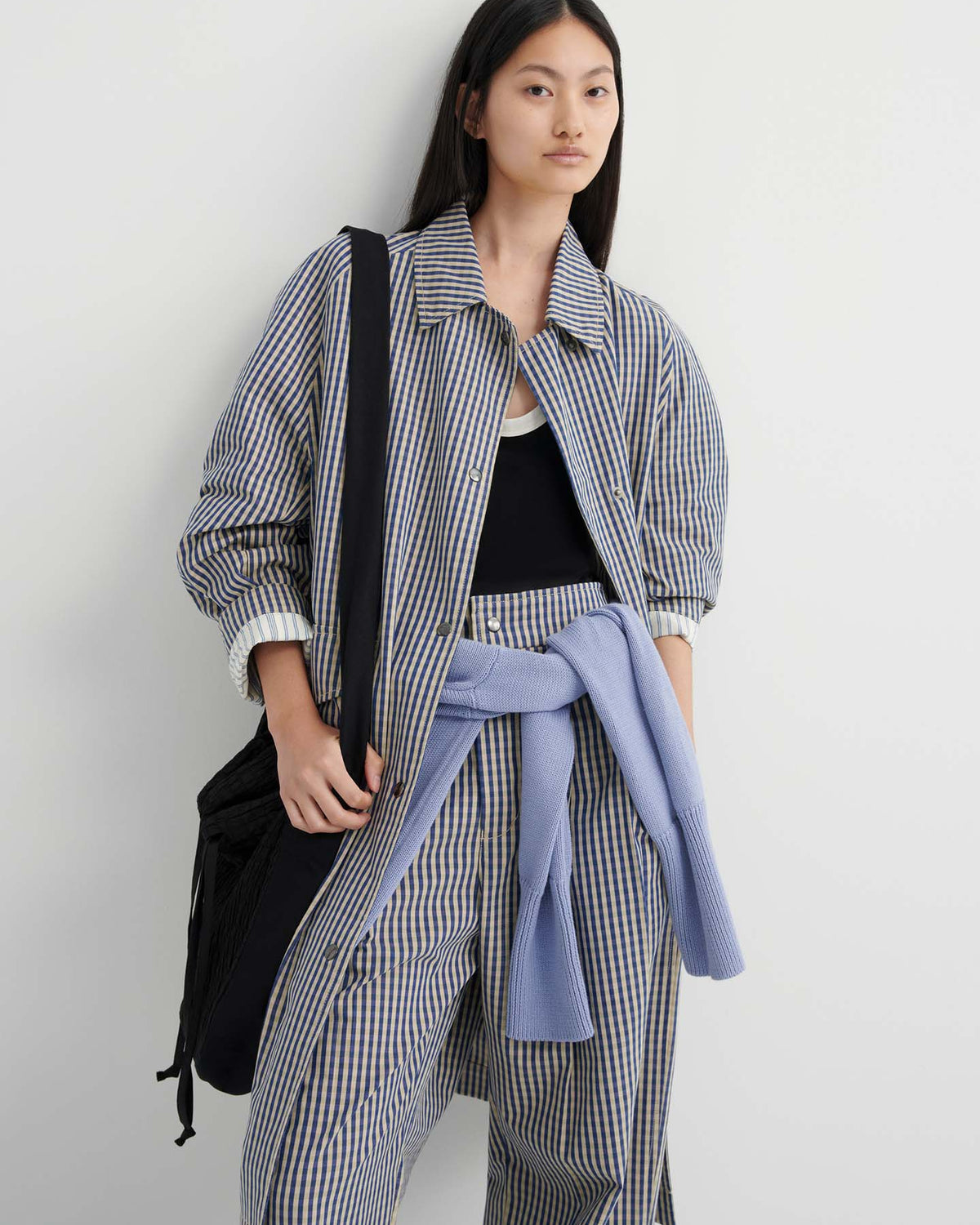 A person stands against a white wall wearing a striped jacket and pants, a black tank top, and a light blue sweater tied at the waist, carrying a black bag styled with the Atelier Trench – Landscape Check for a relaxed trench coat look.