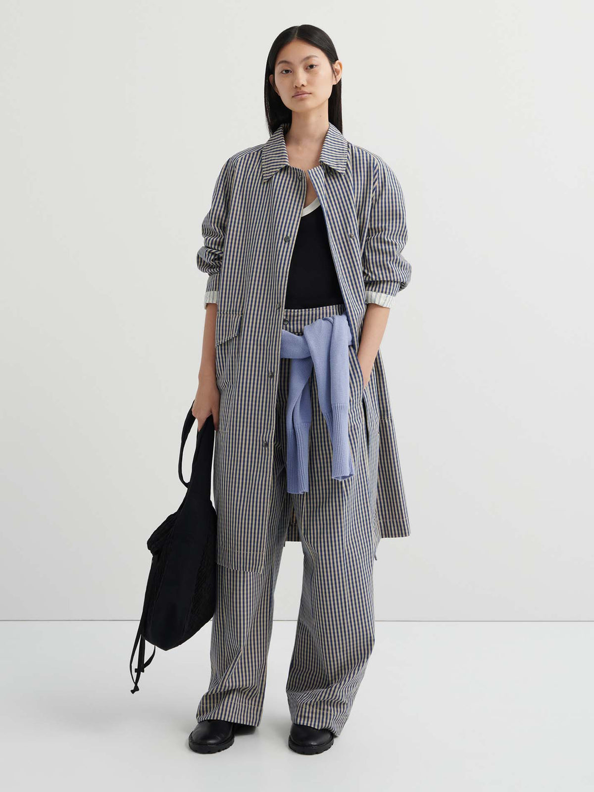A woman stands against a plain background wearing the relaxed fit Atelier Trench – Landscape Check with matching trousers, a light blue sweater tied at her waist, and a black bag for an effortlessly chic look.