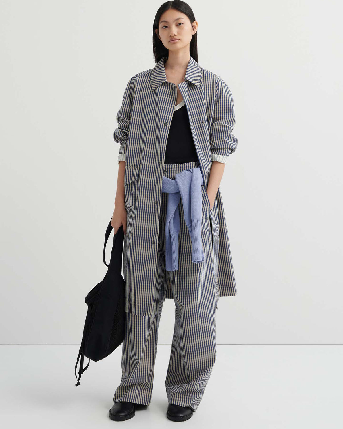A woman stands against a plain background wearing the relaxed fit Atelier Trench – Landscape Check with matching trousers, a light blue sweater tied at her waist, and a black bag for an effortlessly chic look.