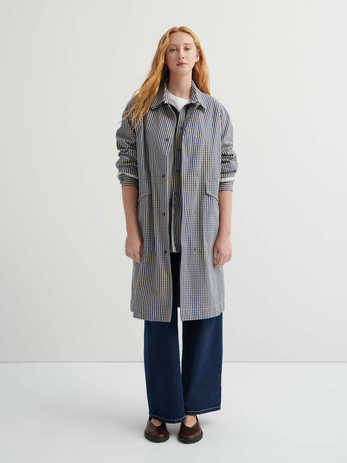 A person stands facing forward, wearing the Atelier Trench – Landscape Check with adjustable snap tabs, wide-leg dark blue jeans, and brown shoes against a plain white background.