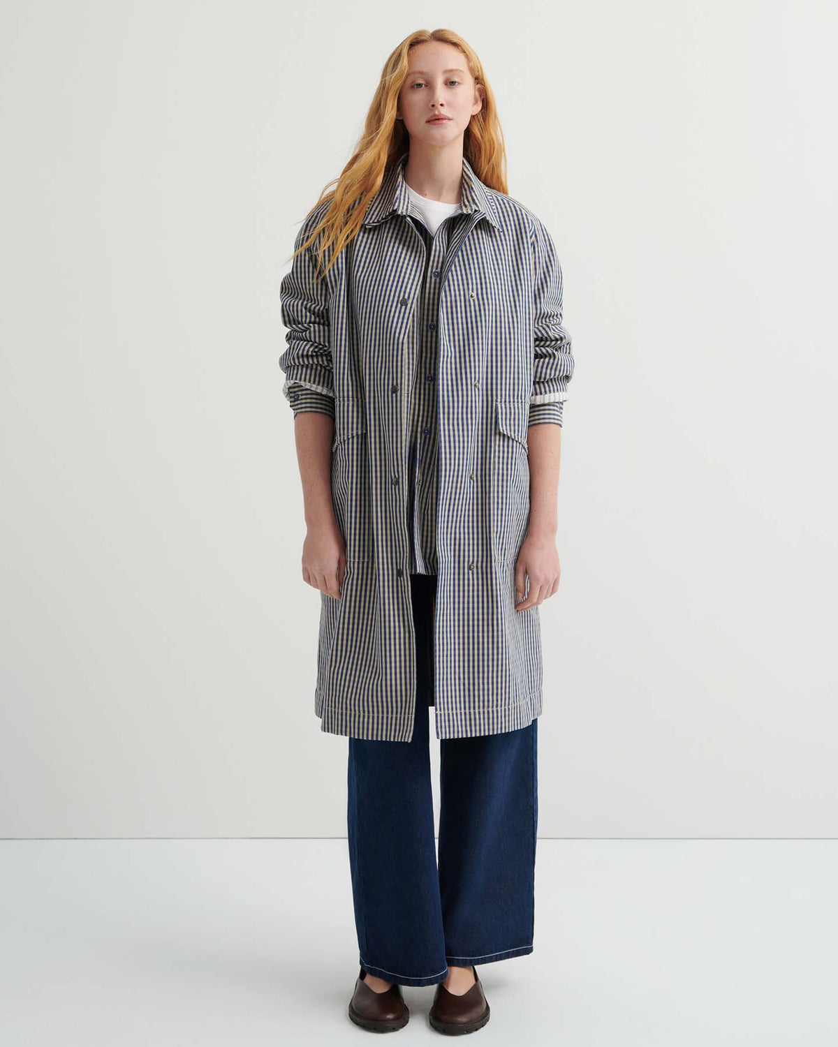 A person stands facing forward, wearing the Atelier Trench – Landscape Check with adjustable snap tabs, wide-leg dark blue jeans, and brown shoes against a plain white background.