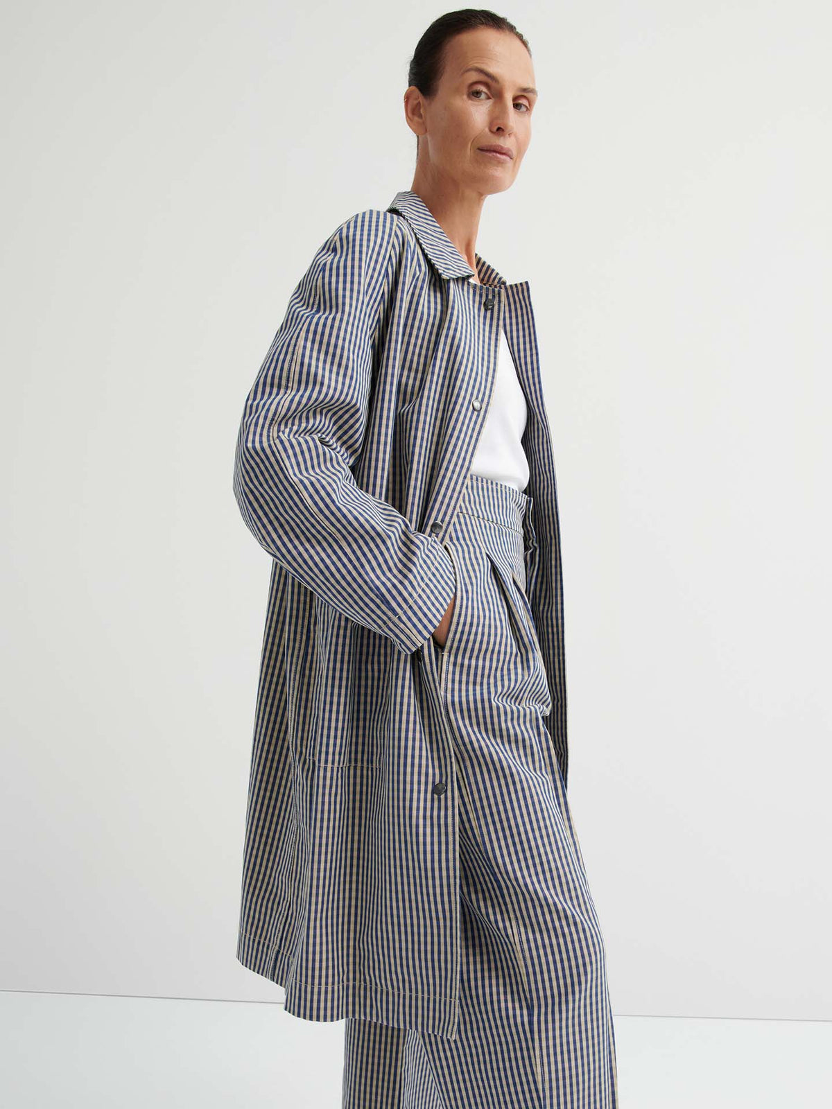 A person wearing the Atelier Trench – Landscape Check, featuring a relaxed fit and adjustable snap tabs, paired with wide-leg pants over a white top, stands against a plain white background.