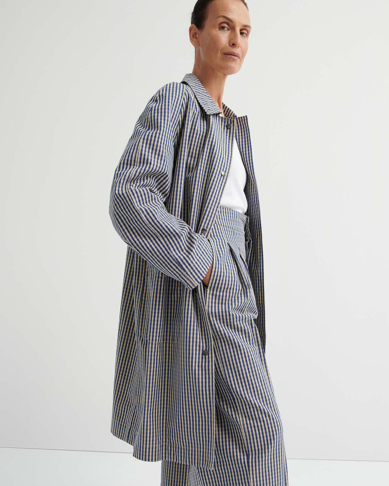 A person wearing the Atelier Trench – Landscape Check, featuring a relaxed fit and adjustable snap tabs, paired with wide-leg pants over a white top, stands against a plain white background.