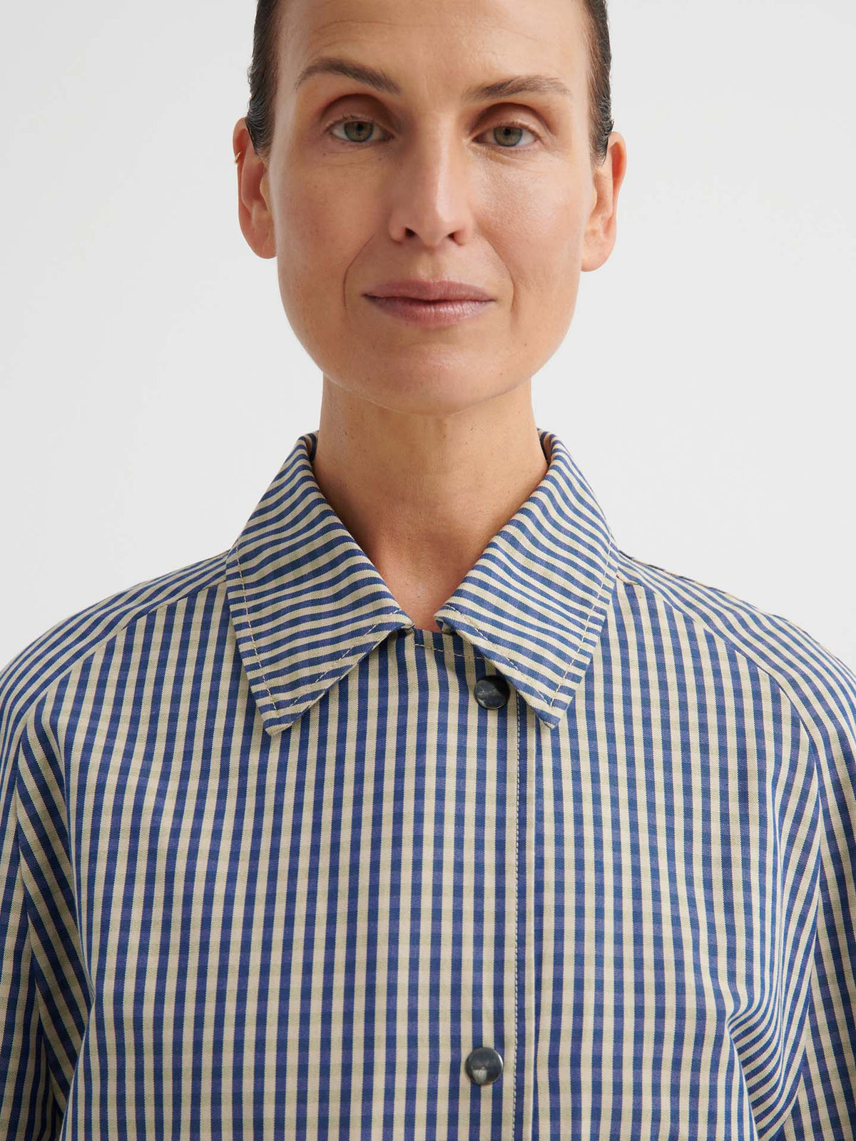 A person stands facing forward against a plain background, wearing a collared blue and white striped shirt layered under the Atelier Trench – Landscape Check with adjustable snap tabs.