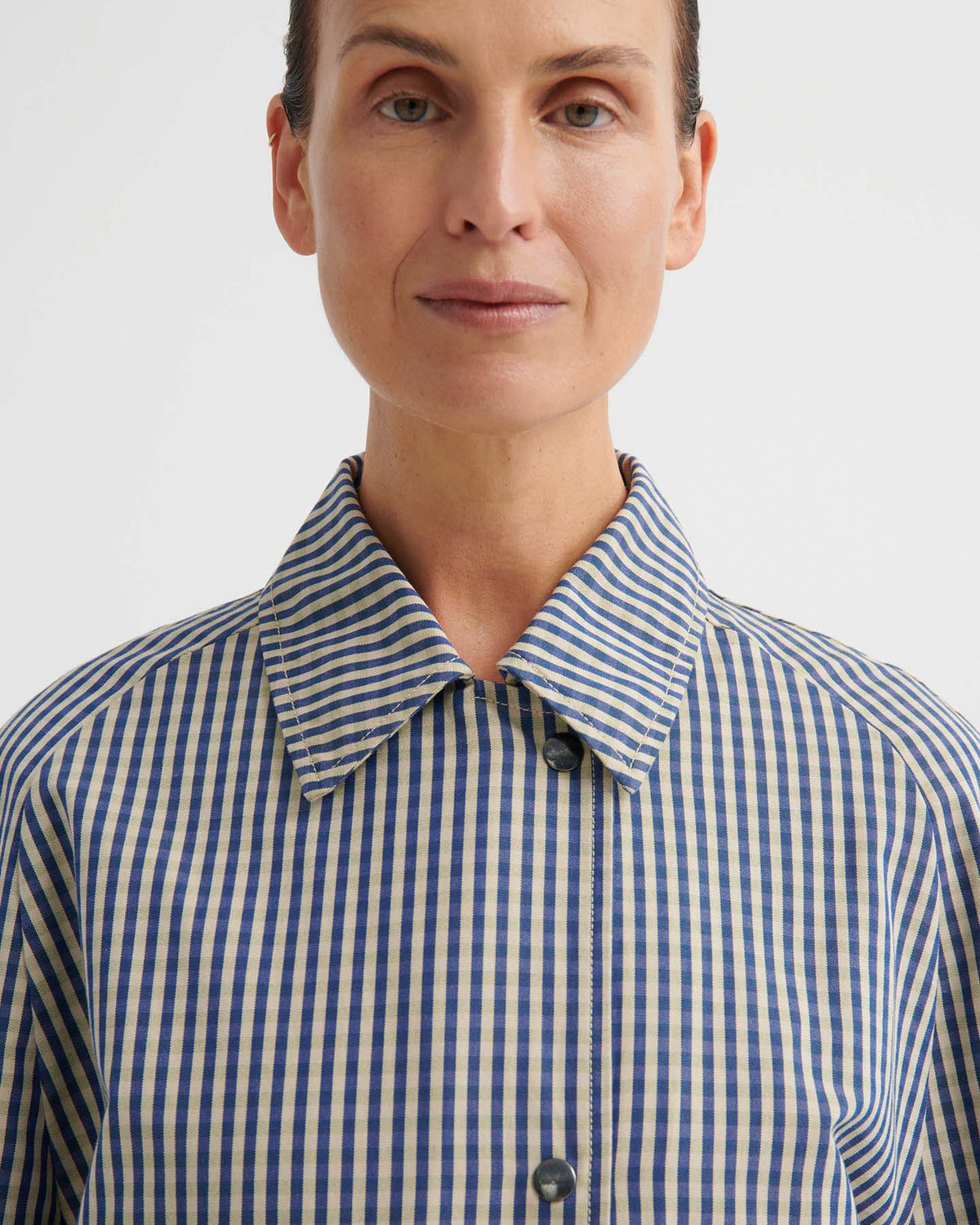 A person stands facing forward against a plain background, wearing a collared blue and white striped shirt layered under the Atelier Trench – Landscape Check with adjustable snap tabs.