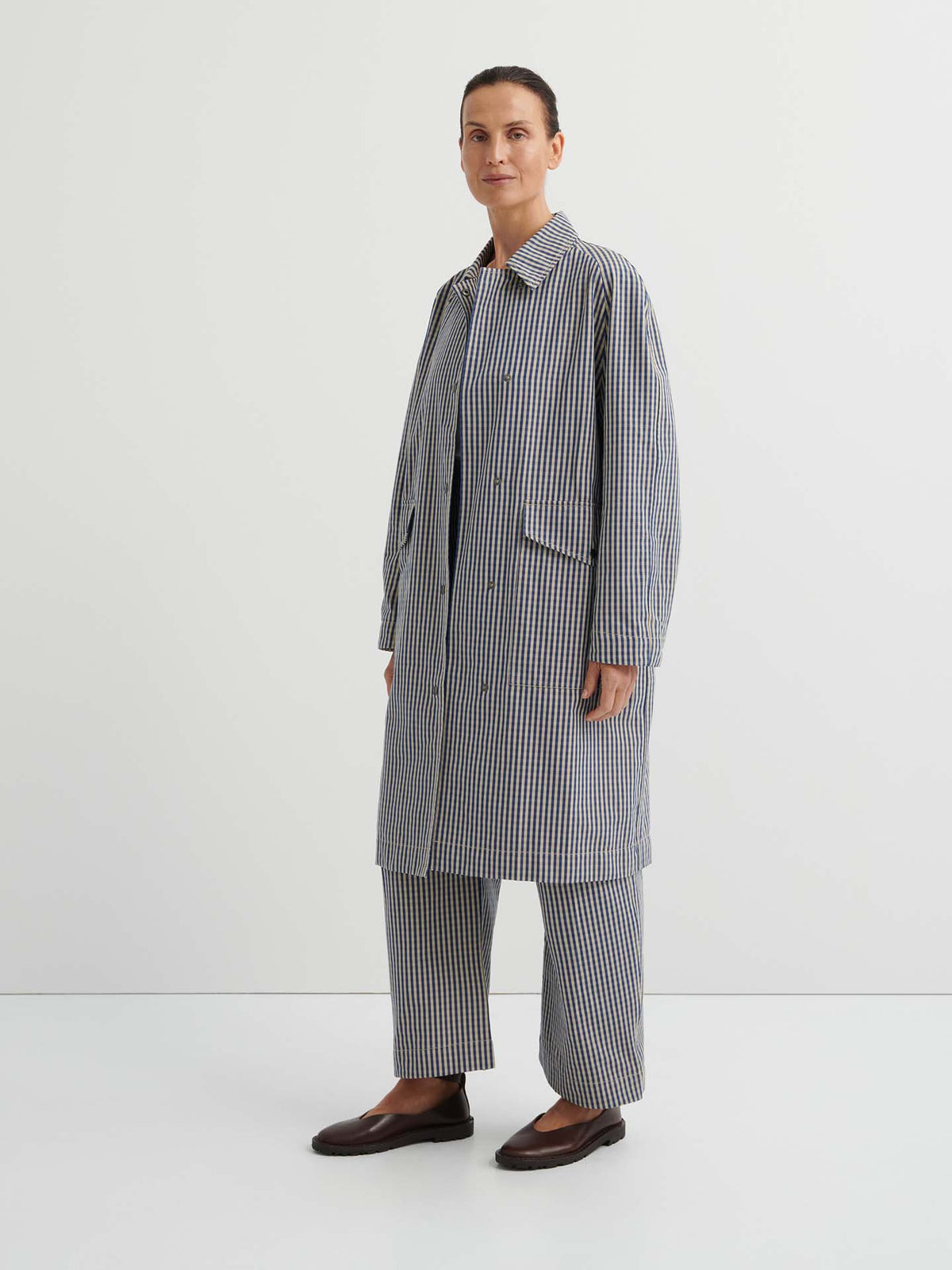 A person stands against a plain background wearing the Atelier Trench – Landscape Check, a relaxed-fit trench coat with adjustable snap tabs, paired with matching wide-leg trousers and dark flat shoes.