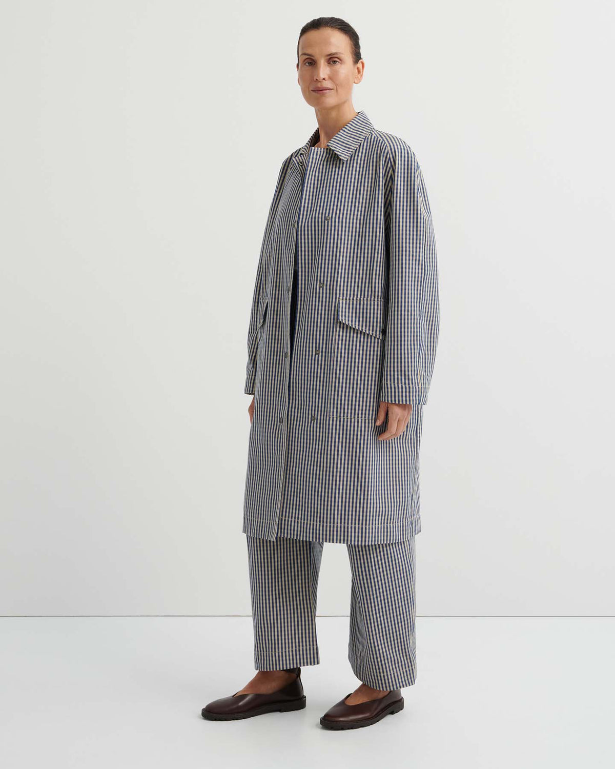 A person stands against a plain background wearing the Atelier Trench – Landscape Check, a relaxed-fit trench coat with adjustable snap tabs, paired with matching wide-leg trousers and dark flat shoes.