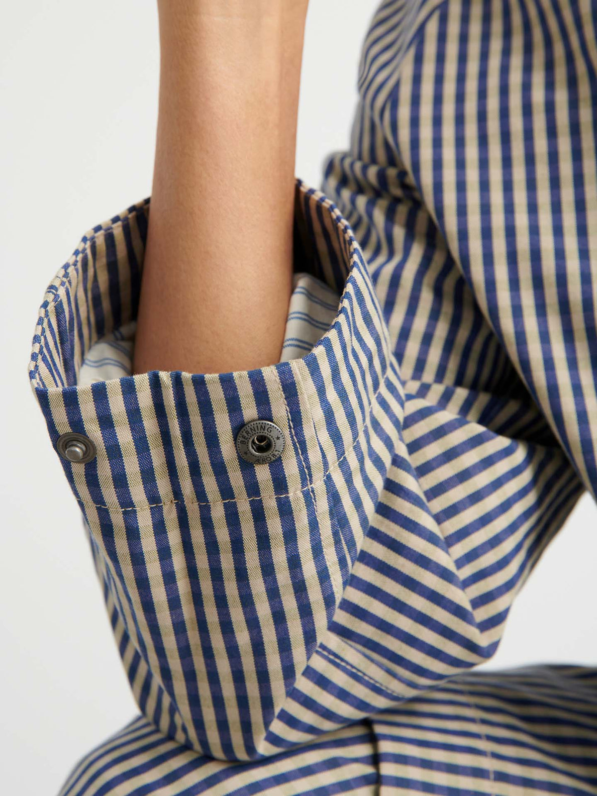 A close-up of a person's arm in a blue and beige striped shirt, the cuff with two visible buttons peeking out from under the Atelier Trench – Landscape Check with adjustable snap tabs.