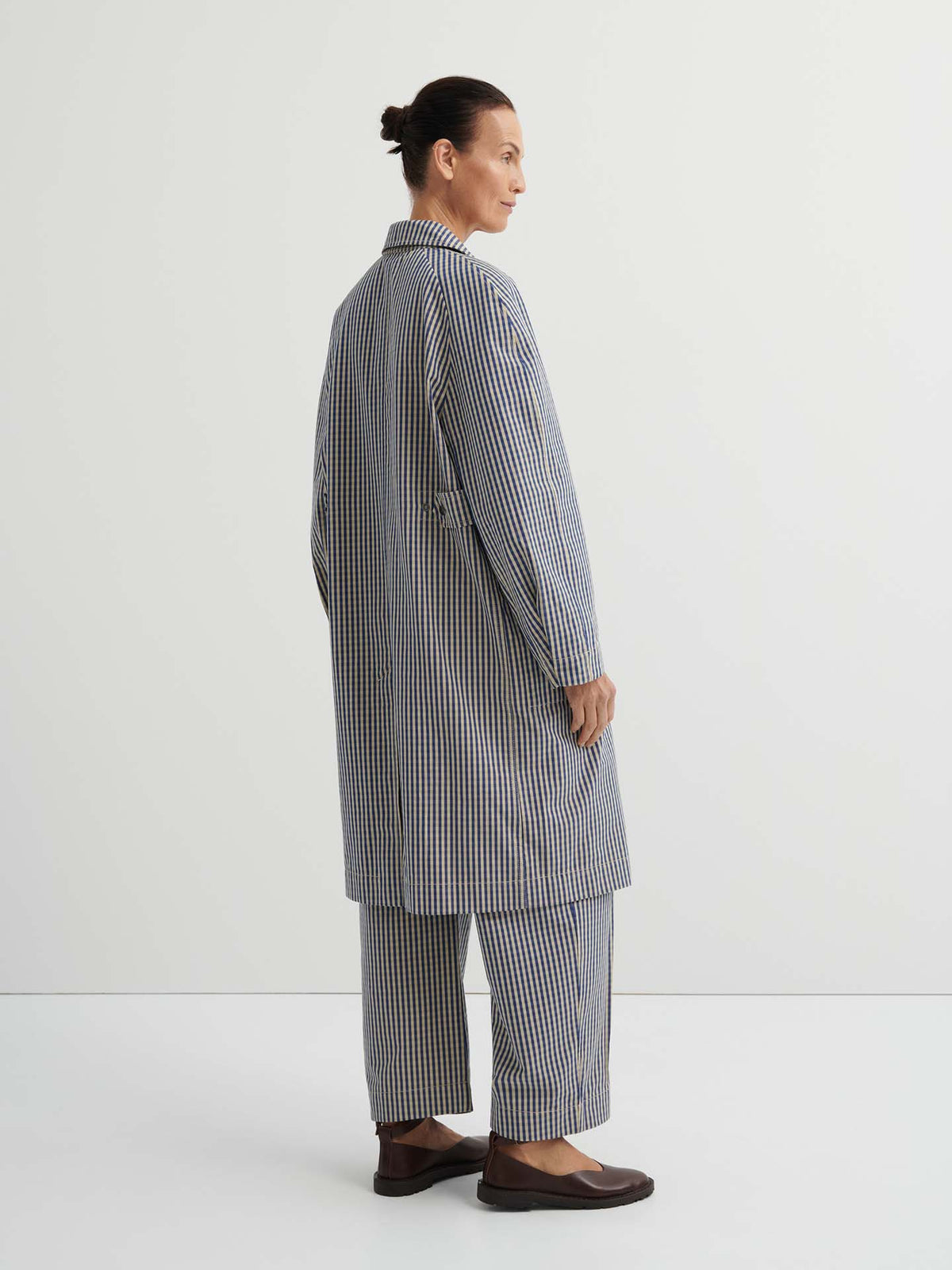 Person standing sideways in the Atelier Trench – Landscape Check, styled with matching checkered pants featuring adjustable snap tabs and brown shoes, set against a plain white background.