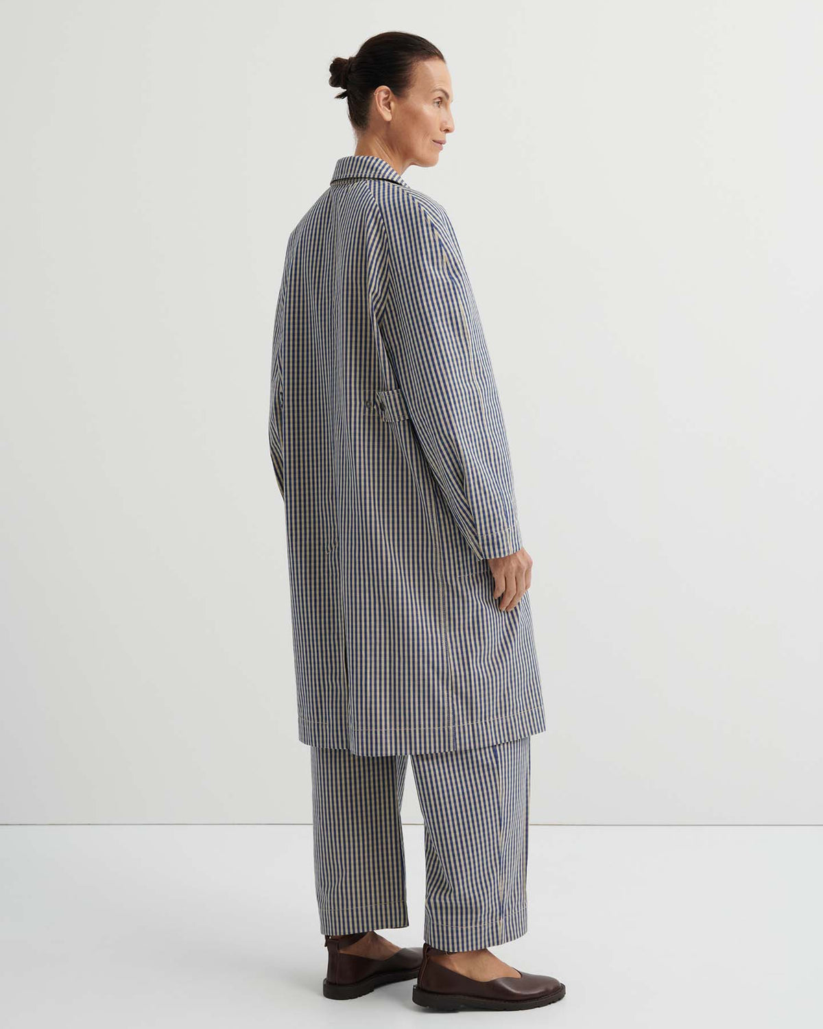 Person standing sideways in the Atelier Trench – Landscape Check, styled with matching checkered pants featuring adjustable snap tabs and brown shoes, set against a plain white background.