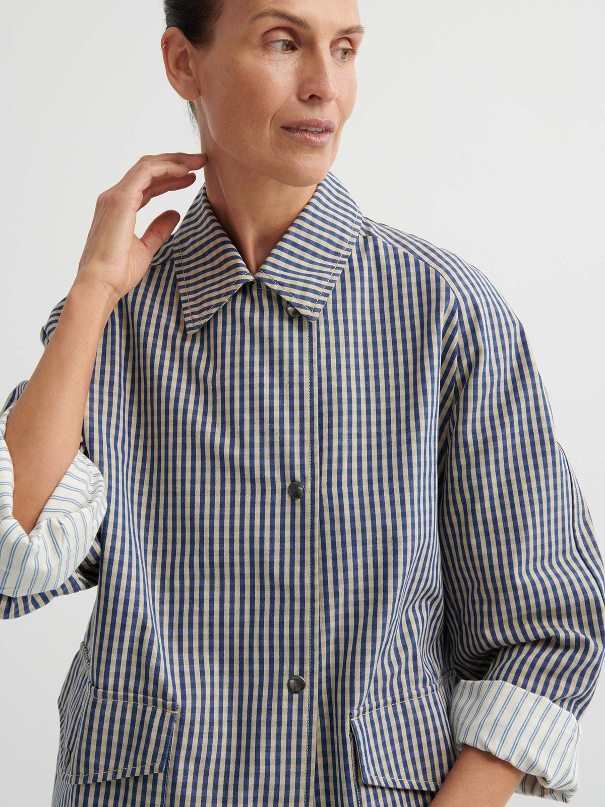 A woman in an Atelier Trench – Landscape Check with adjustable snap tabs and rolled sleeves looks to the side against a plain background.