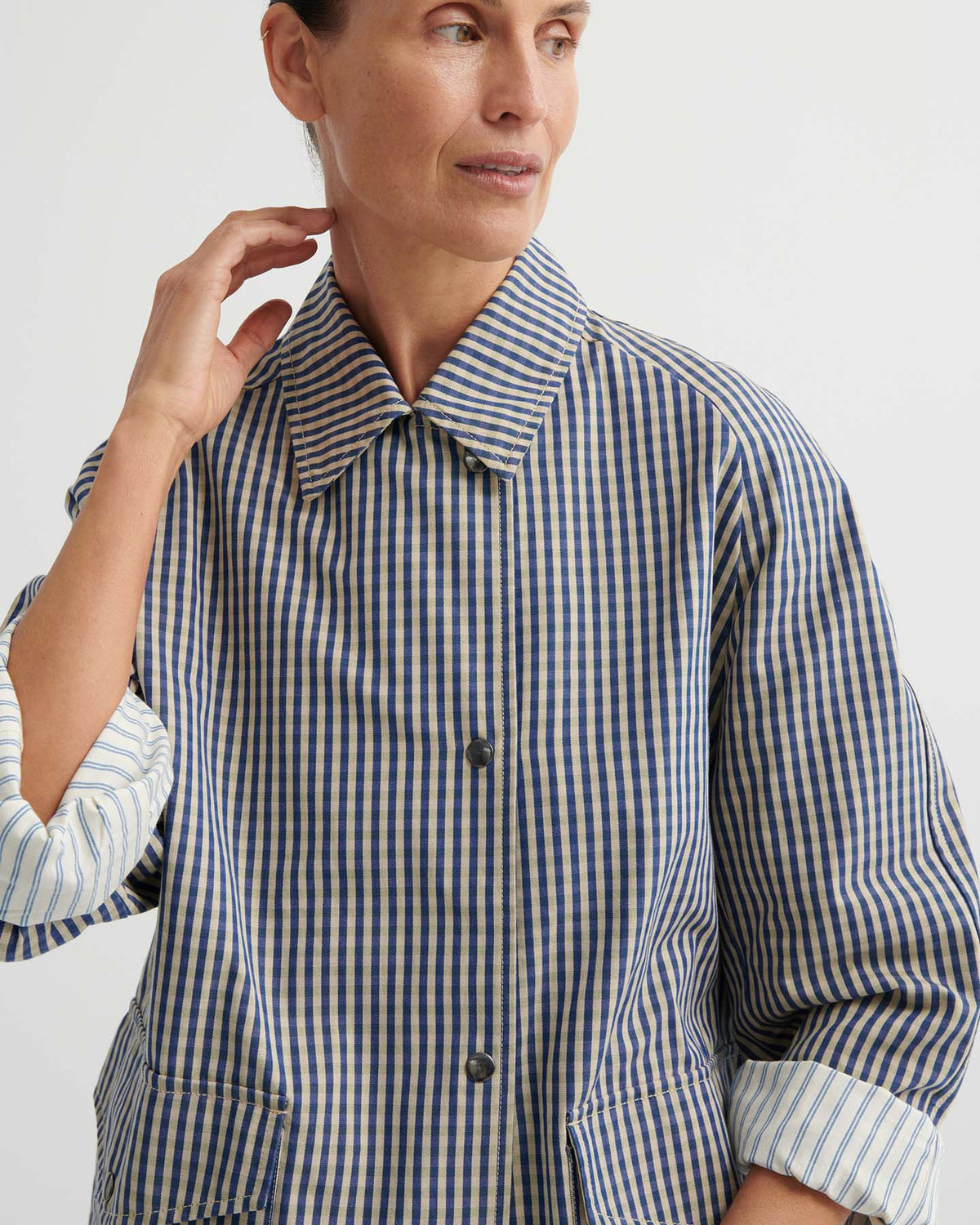 A woman in an Atelier Trench – Landscape Check with adjustable snap tabs and rolled sleeves looks to the side against a plain background.