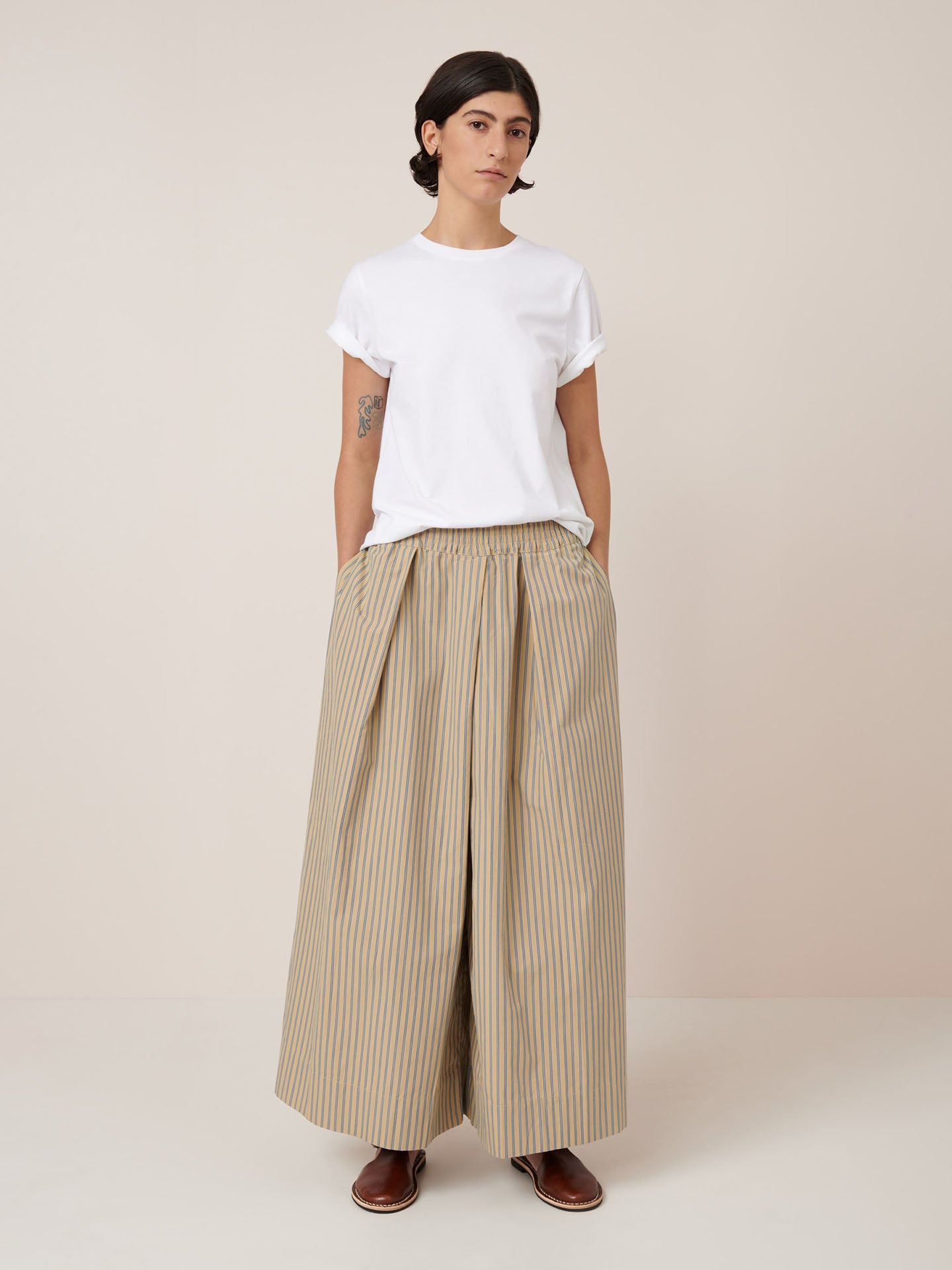 A person stands against a plain background, wearing Kowtow’s Athena Pant – Double Pinstripe with an elastic true-waist and wide legs, paired with a white tee, brown shoes, and hands in pockets.
