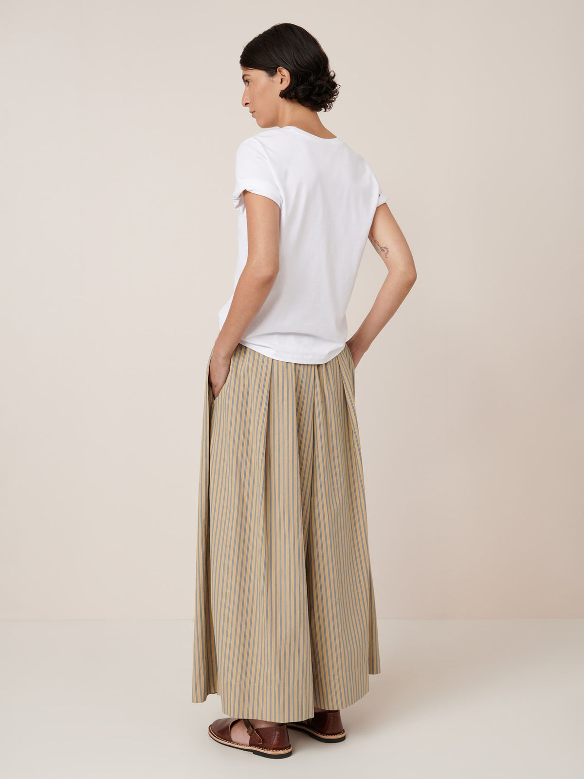 A person faces away from the camera, hands in pockets, wearing a white short-sleeve shirt and Kowtow’s Athena Pant – Double Pinstripe: relaxed fit beige and yellow striped pants with an elastic true waist, paired with brown sandals.
